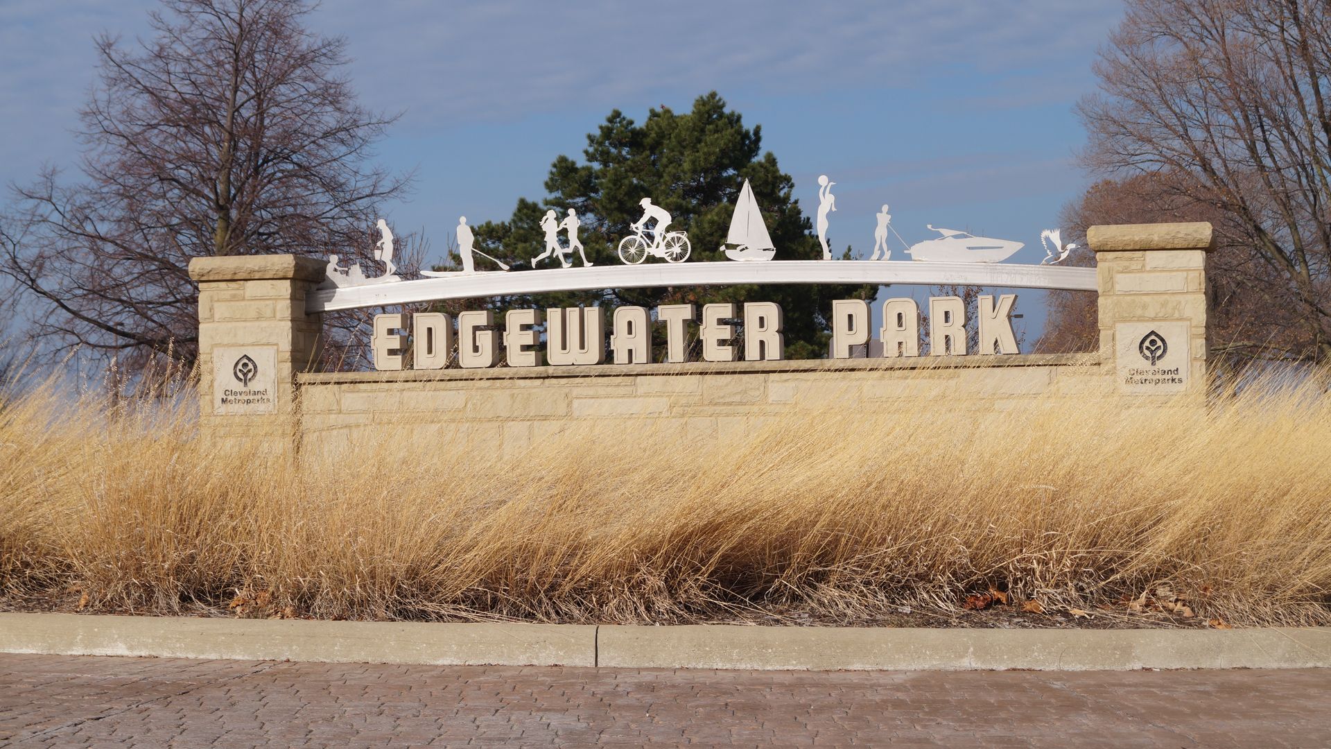 Edgewater Park entrance sign with white silhouettes of people biking, walking, sailing, and a boat atop a curved arch. Beige stone columns, Cleveland Metroparks logos, blue sky, bare trees, and tall dry grass in foreground.