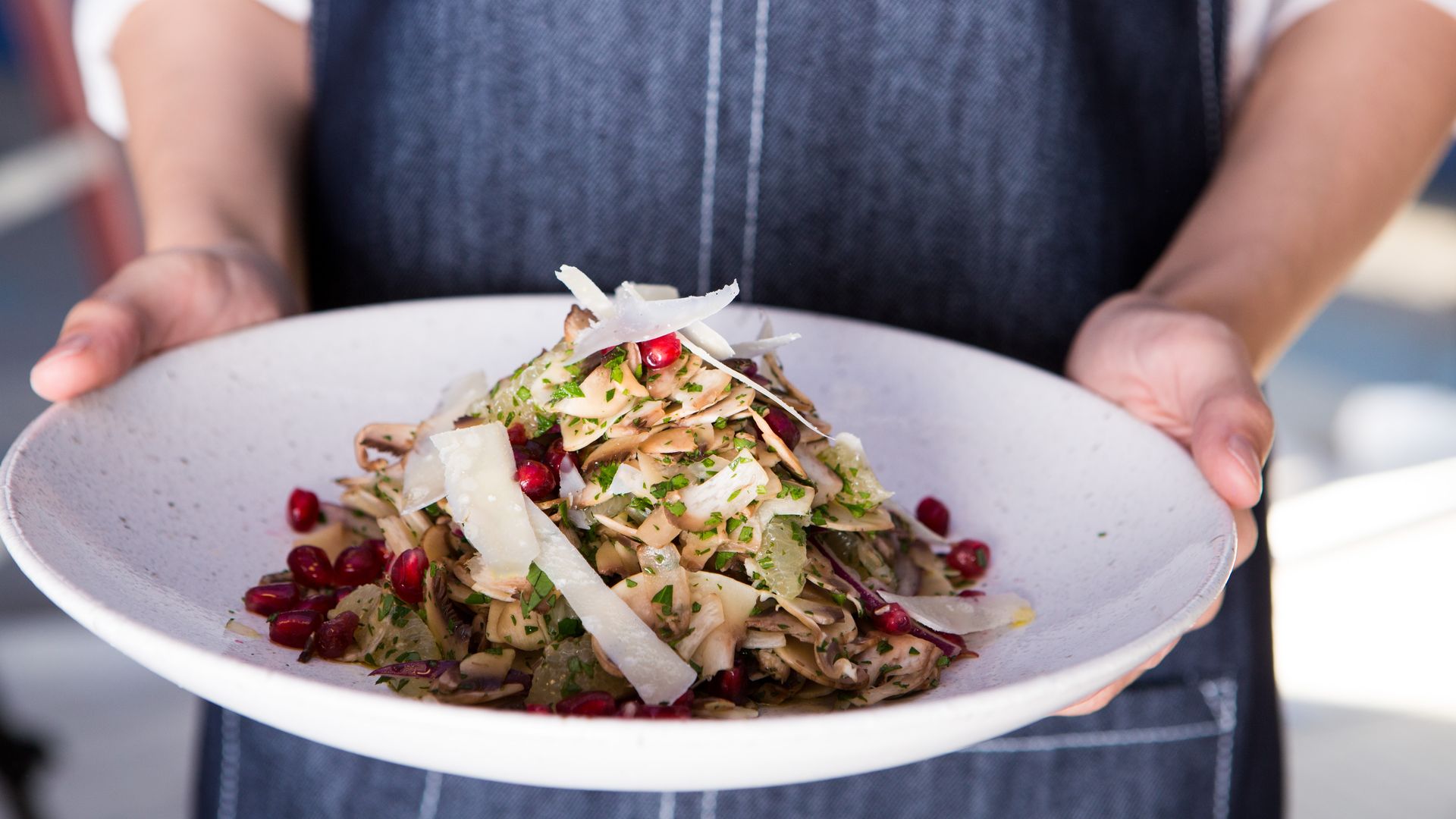 A person in a dark denim apron holds a white plate with a colorful white mushroom salad from Clementine topped with pomegranate seeds and shaved Pecorino.