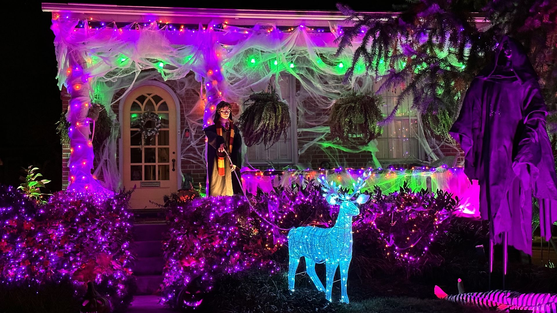 Nighttime Halloween porch with purple, pink, and green lights, white spiderweb netting over the railing, a cloaked figure on the steps, and a glowing blue deer statue in the garden.