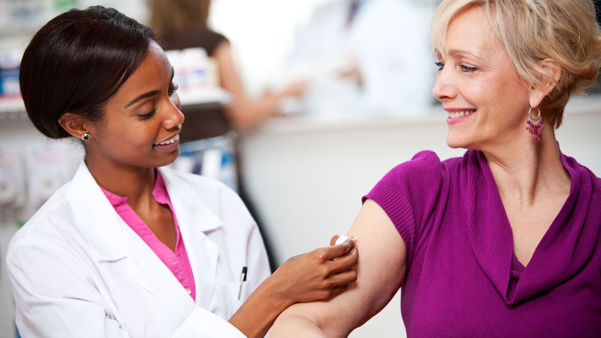 A female healthcare professional in a white coat and pink shirt applies a bandage on the upper arm of a smiling woman wearing a purple top and earrings in a clinical setting.