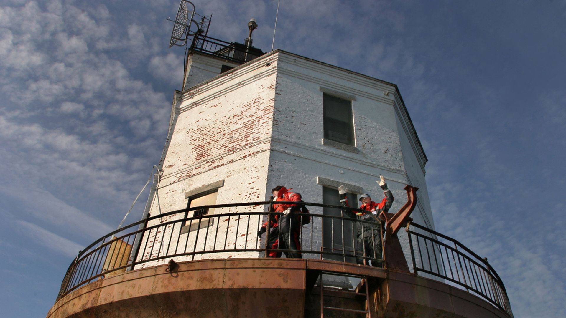 A view of a lighthouse from below with people waving from the wraparound porch-type thing. The lighthouse behind them is white with faded red brick peeking through