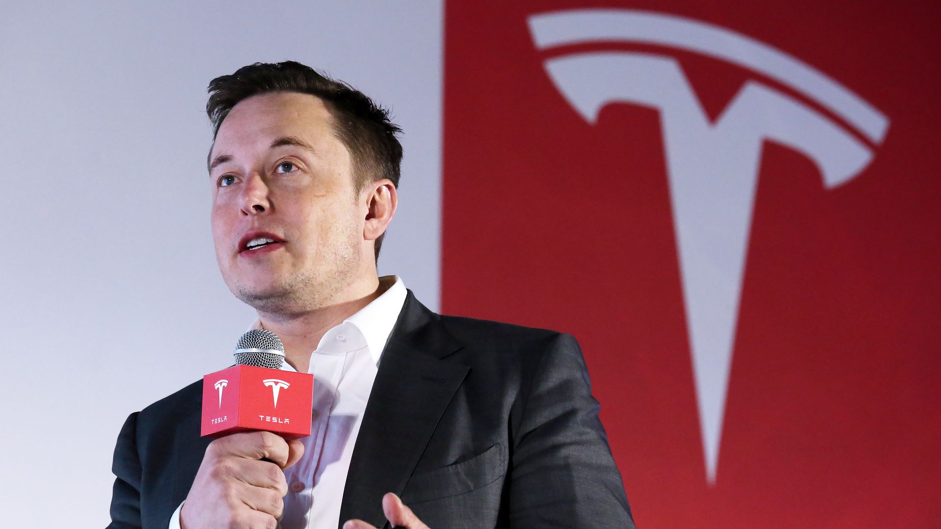 Elon Musk, in a dark suit and white shirt, speaks into a microphone with a red Tesla logo cover, with a large red and white Tesla logo in the background.