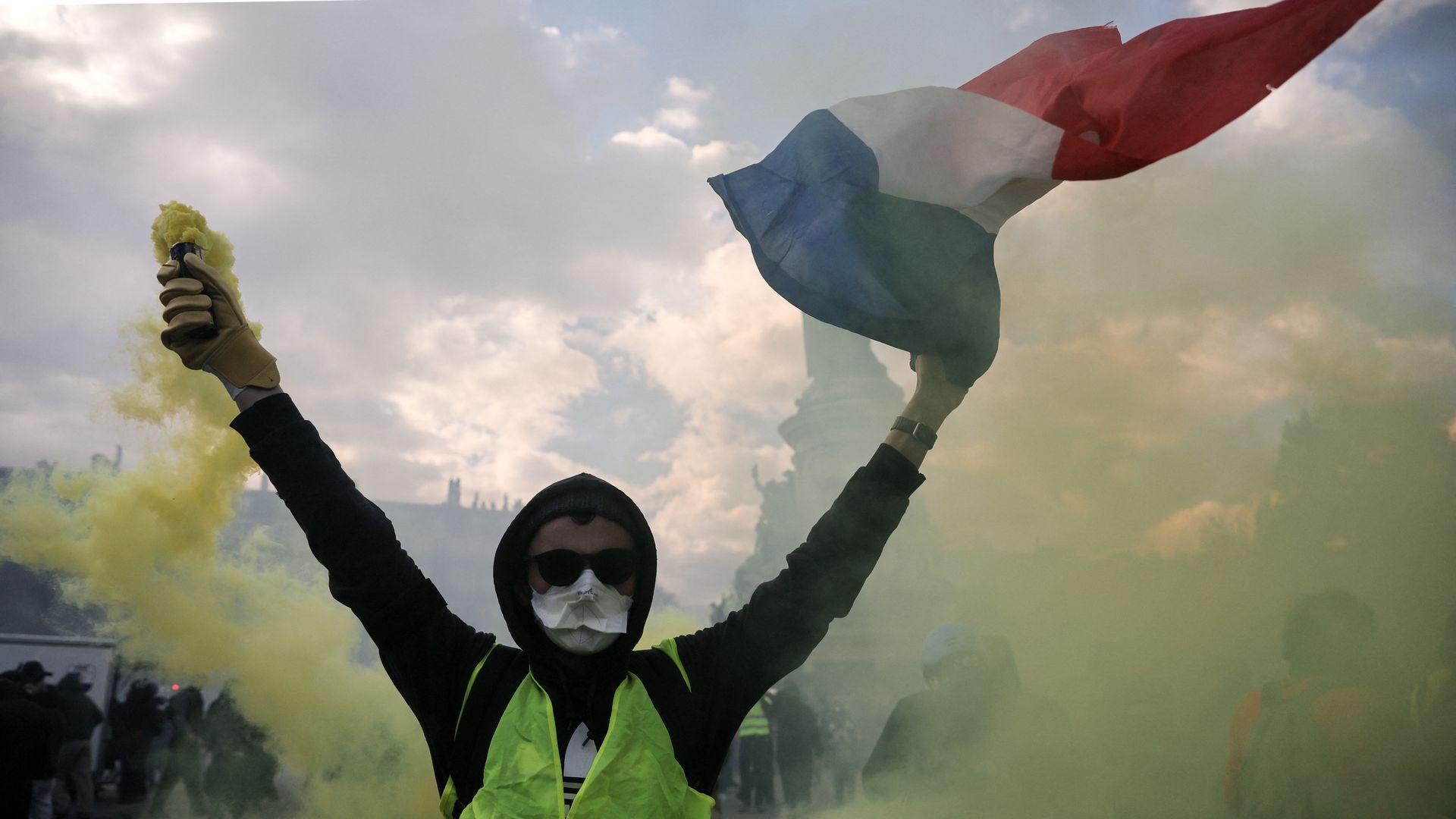 A person wearing a dark hoodie, a yellow vest, a white mask and black sunglasses holds a flare putting off yellow smoke in one hand and a French flag in the other during a protest.