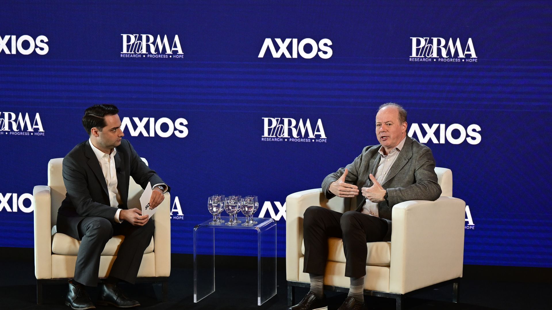 Two men sit in white armchairs on a stage with a blue backdrop featuring Axios and PhRMA logos. A glass side table with several glasses sits between them as they converse.