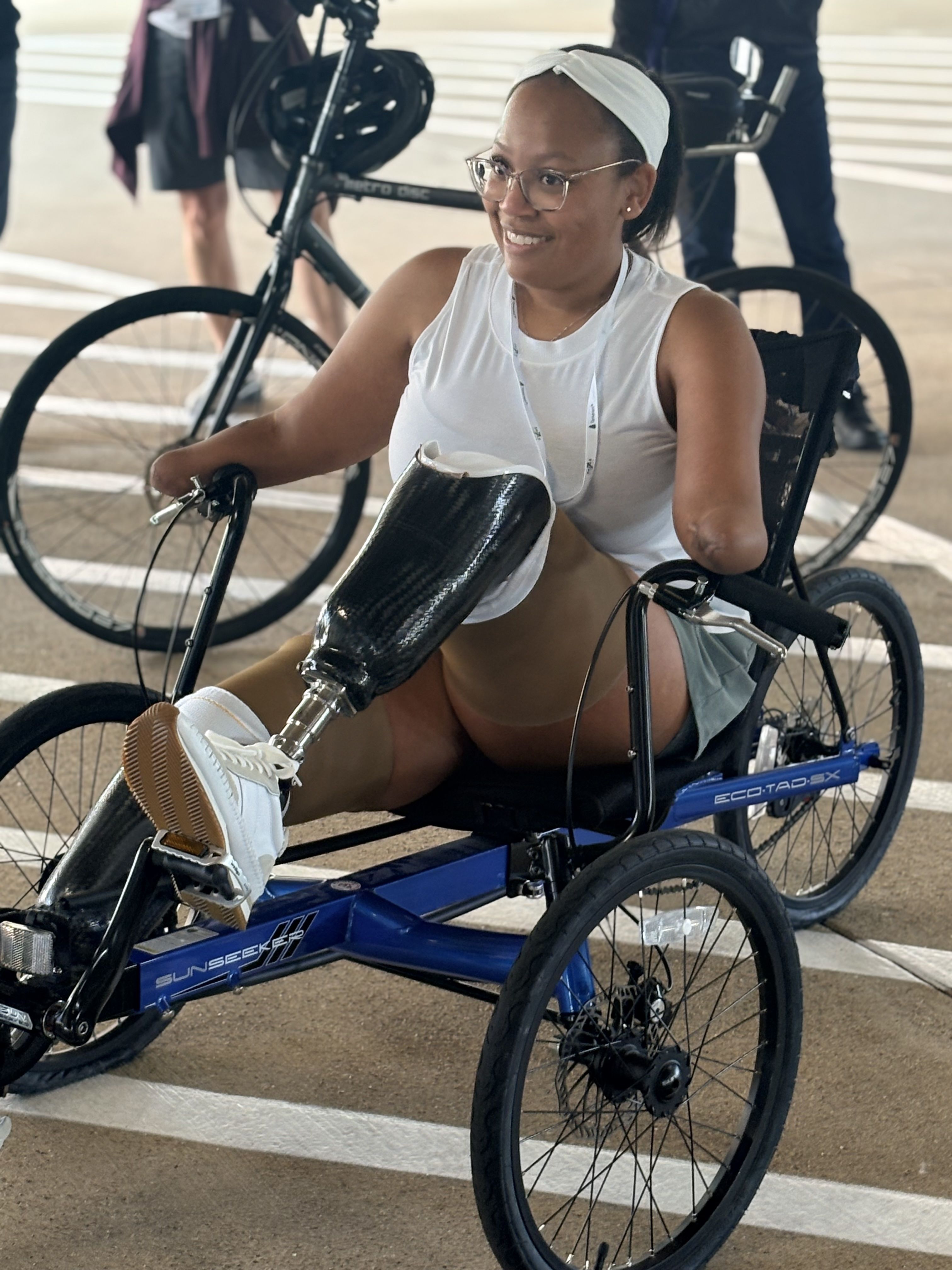 Smiling woman with prosthetic leg and arm riding a blue recumbent tricycle indoors, wearing glasses, white sleeveless top, and white sneakers on the pedal.