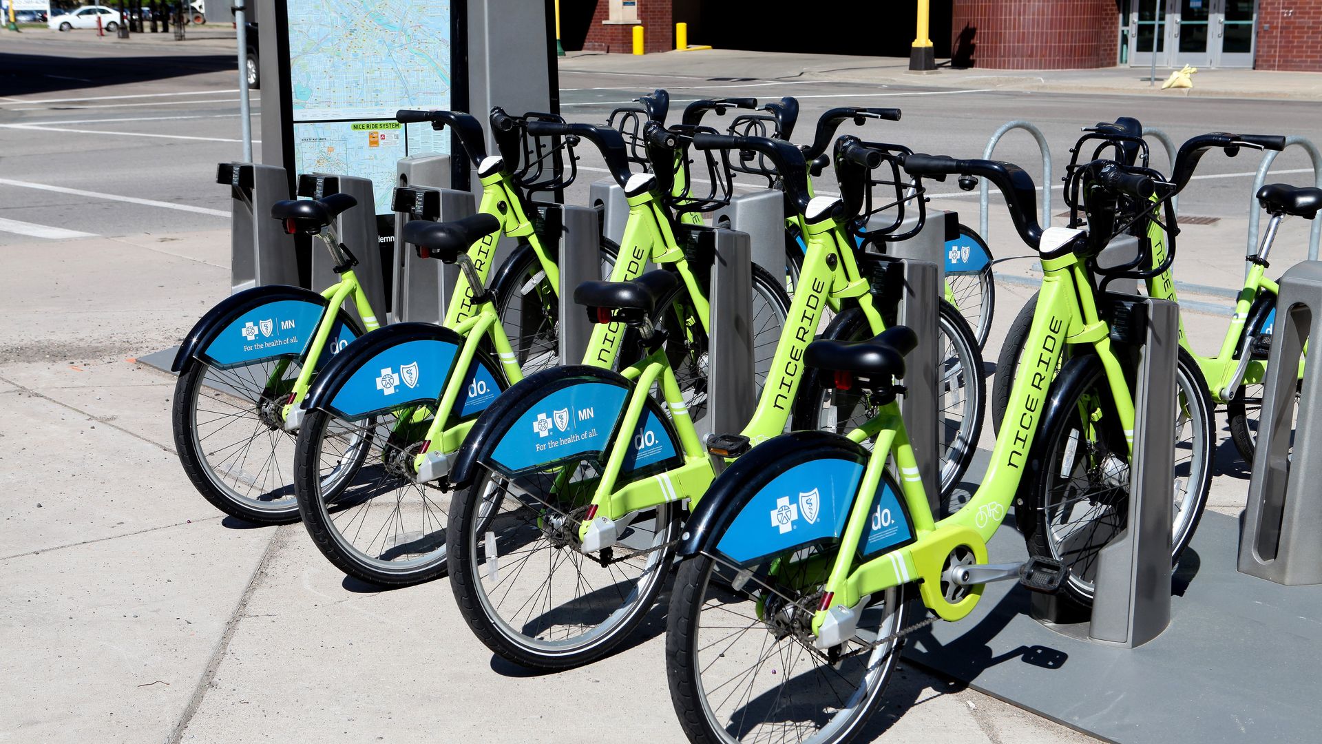 Row of lime-green bike-share bicycles docked at a station on a city sidewalk, with blue wheel covers and black handlebars; a map kiosk is visible in the background.