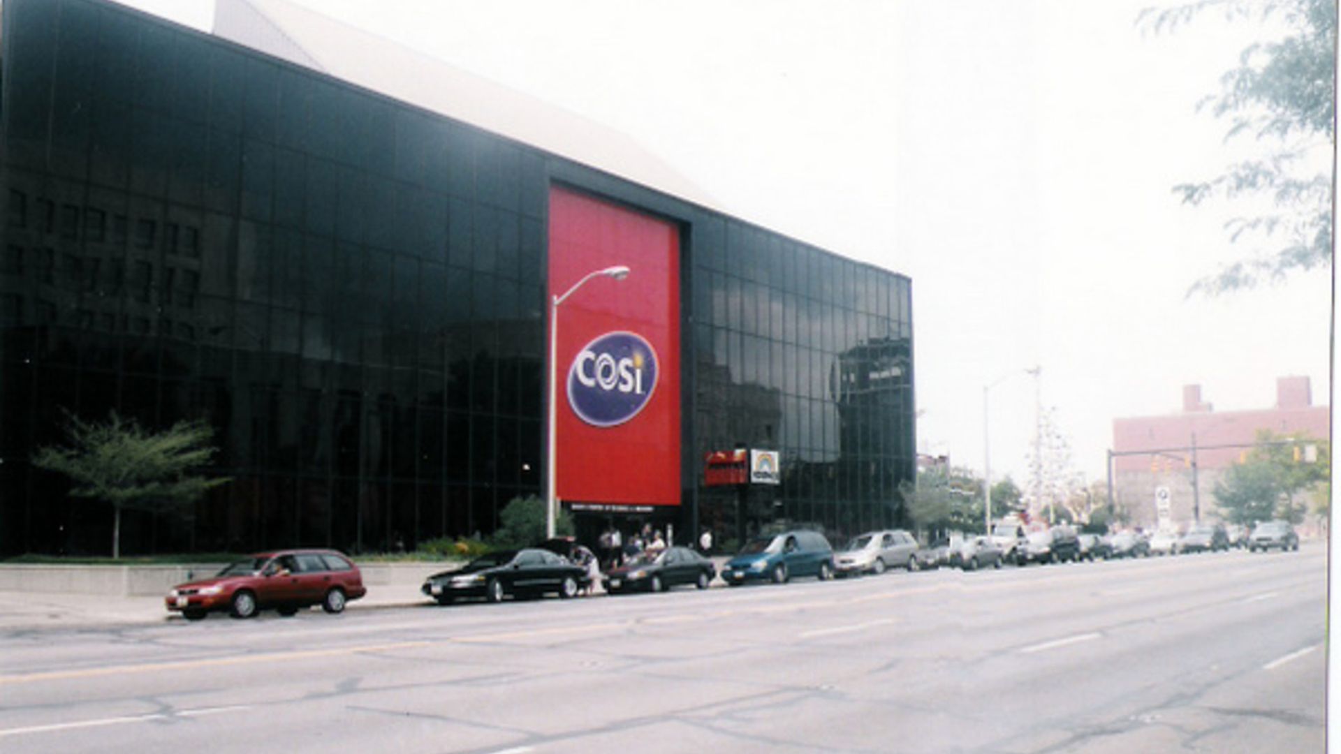 Old, grainy film photo with a sreet view of a large black building with a tall red section featuring a blue oval sign reading "COSI". Several cars are parked along the street in front of the building.