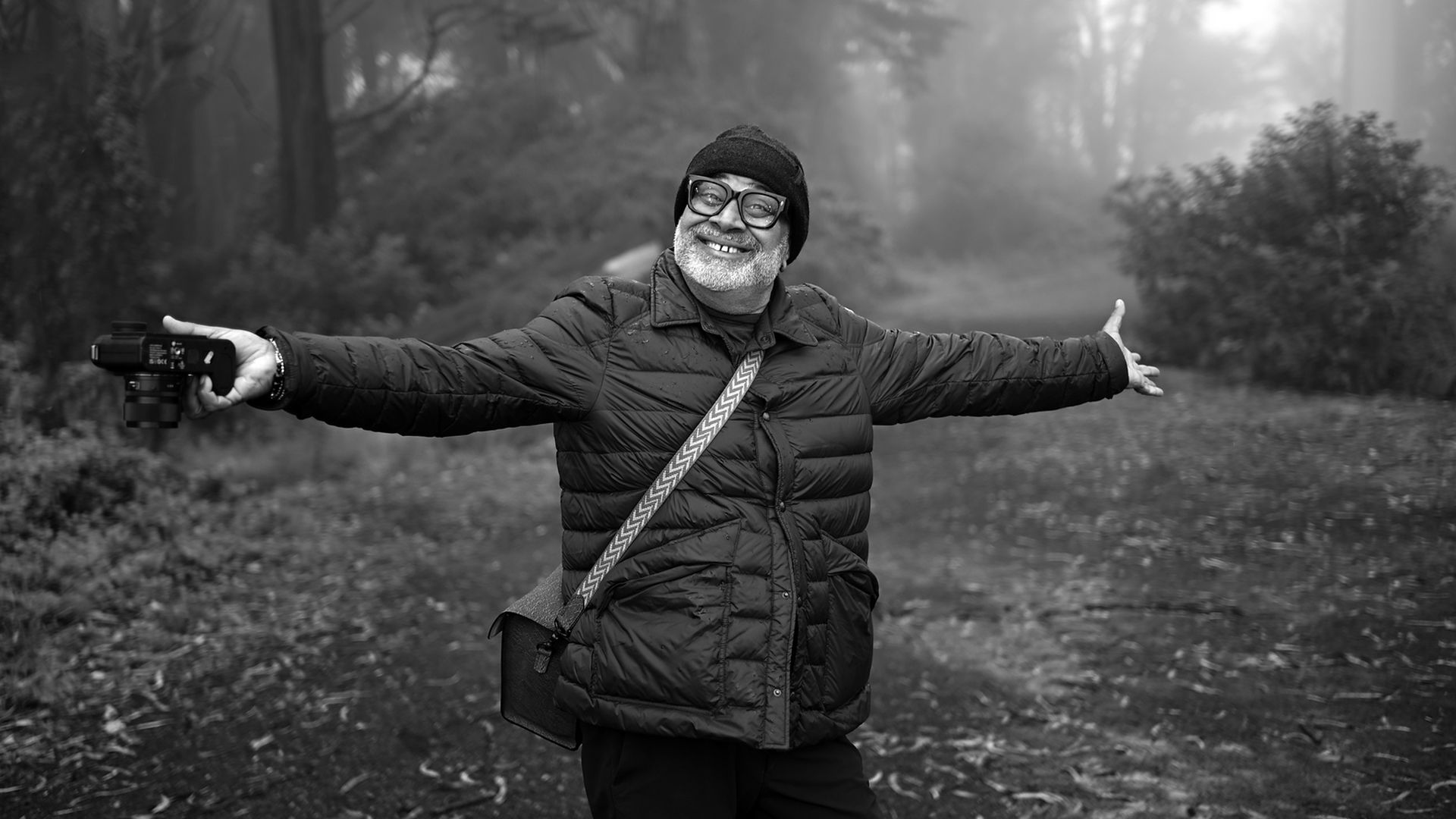 Black-and-white photo of an older man with glasses and a beanie, wearing a quilted jacket and crossbody strap, arms outstretched while holding a camera in a park.