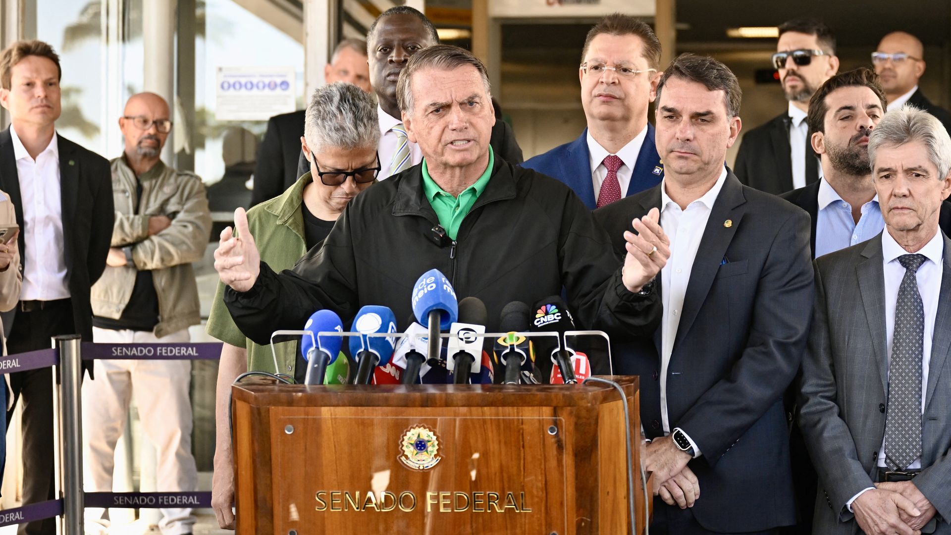 Former Brazilian President Jair Bolsonaro, wearing a black jacket and green shirt, speaks at a wooden podium labeled "Senado Federal" with multiple microphones, surrounded by men in suits and casual clothes outside a building.