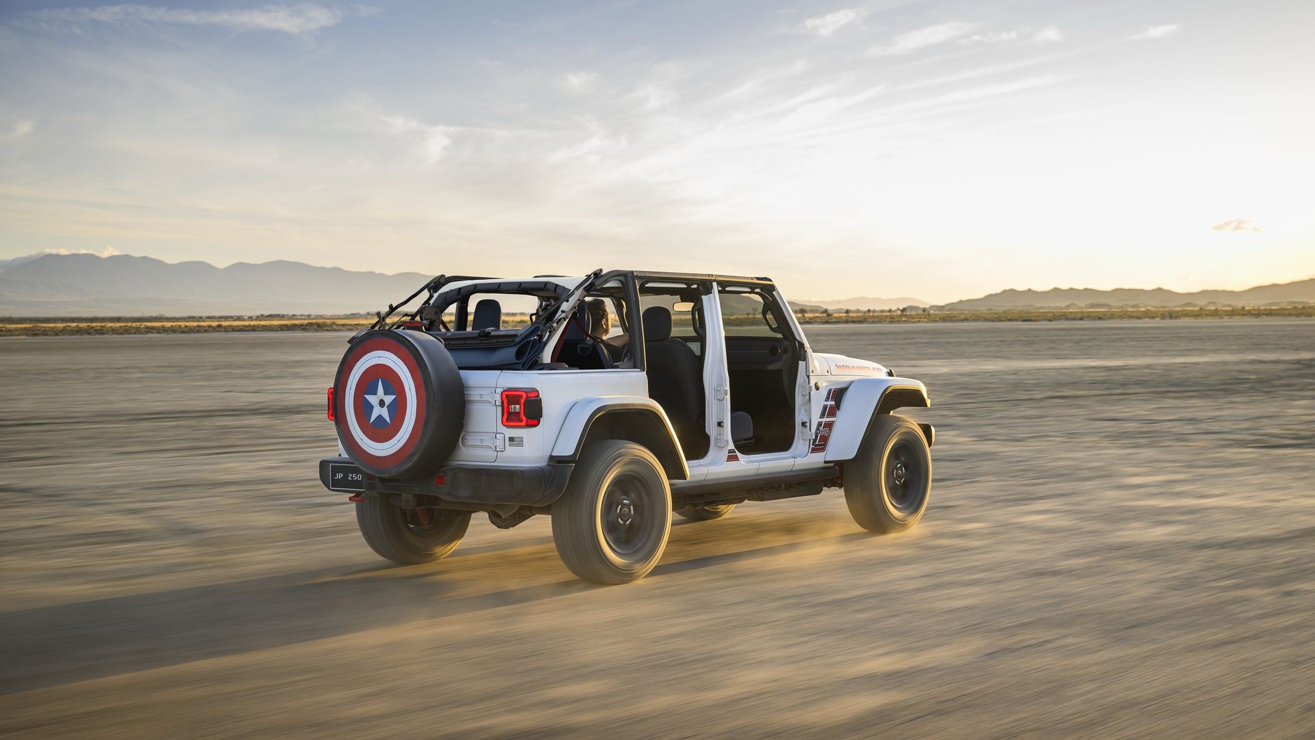 White Jeep Wrangler with open doors and a spare tire cover displaying Captain America's red white and blue shield, speeds across a dusty desert at sunset with mountains in the distance. 