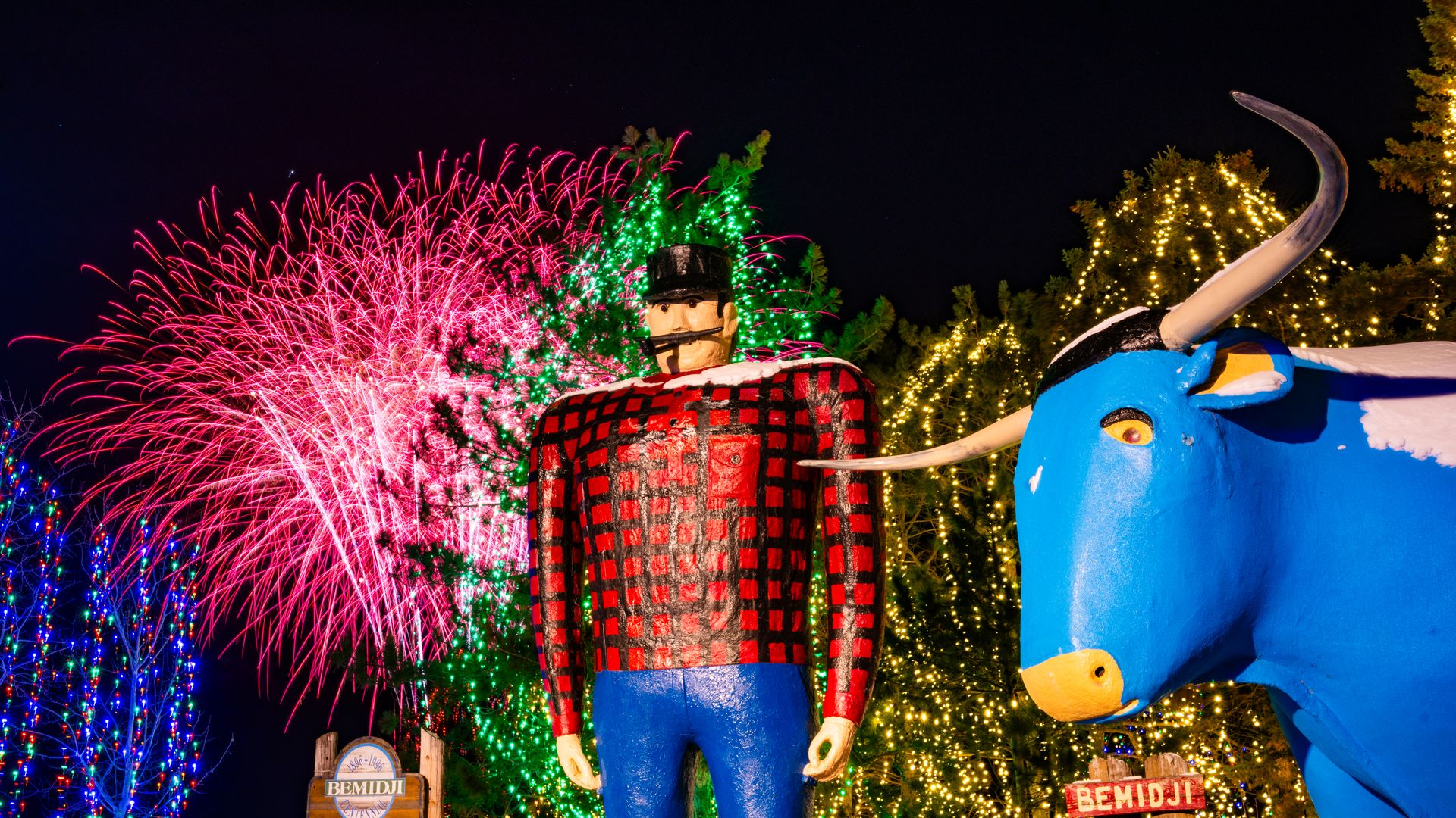 Nighttime festival float: giant red-and-black plaid figure with a black top hat, blue pants, and a horned blue animal beside string lights and pink and green fireworks exploding in the dark sky.