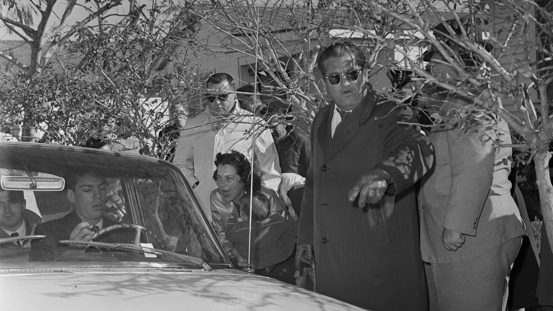 Image shows a black and white photo of people yelling at a man in the car.