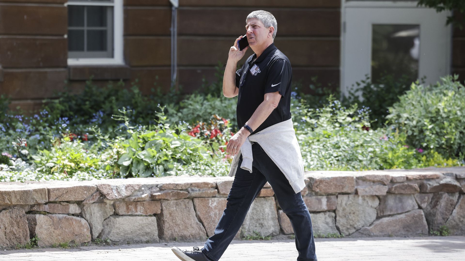 A person in a black polo and dark jeans walks on a sunny sidewalk, talking on a cell phone; a light gray sweatshirt is tied around his waist, with a stone wall and flowering garden behind him.
