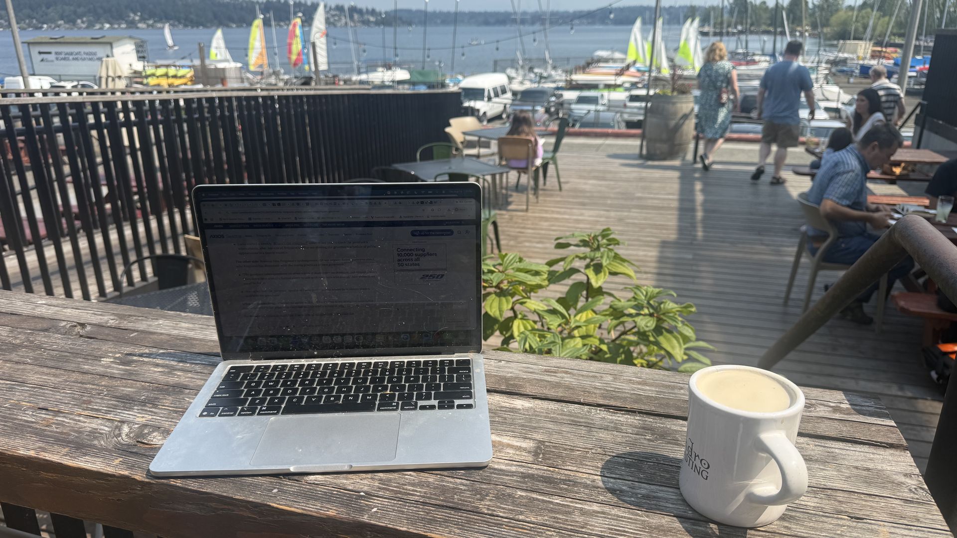 Laptop on a weathered wooden table at a sunny waterfront café, with a white mug in the foreground. In the background, a marina full of sailboats and people strolling along the dock.