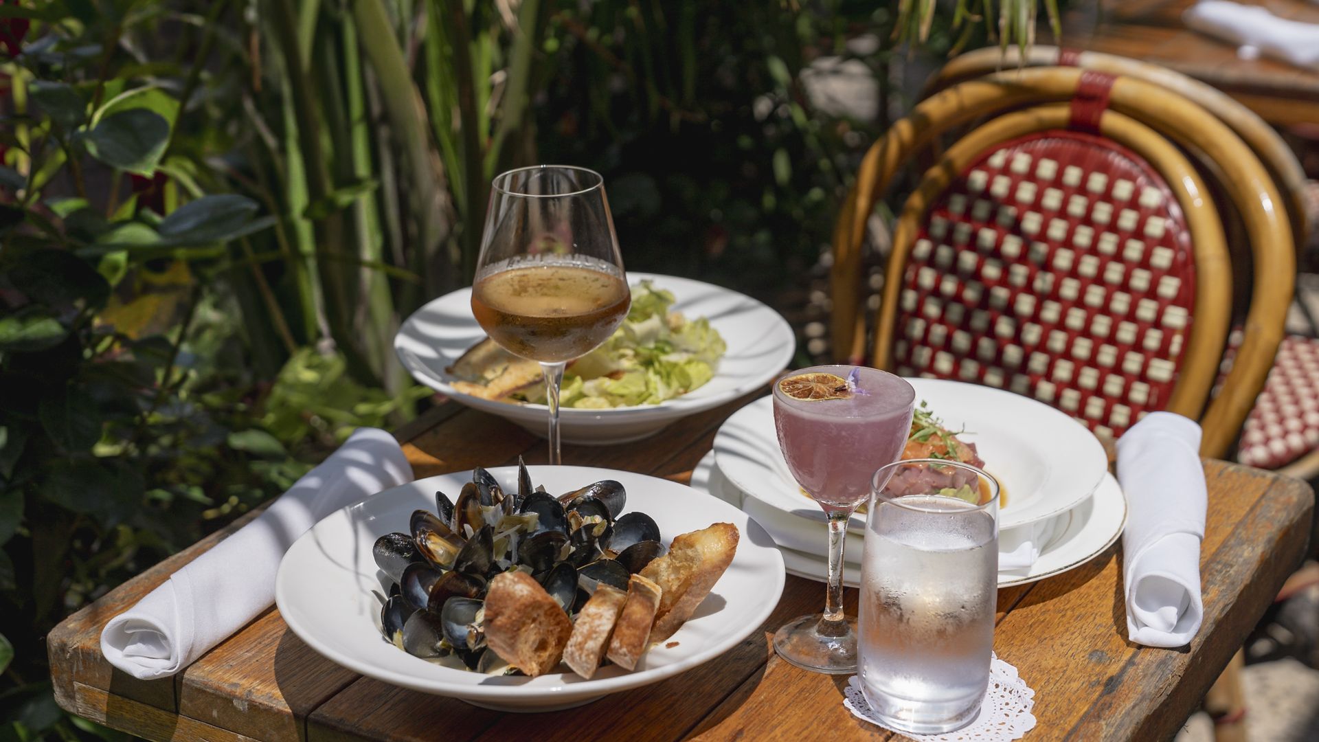 A white plate of mussels with a glass of wine, pink cocktail, water and two other dishes of food on a wood table.