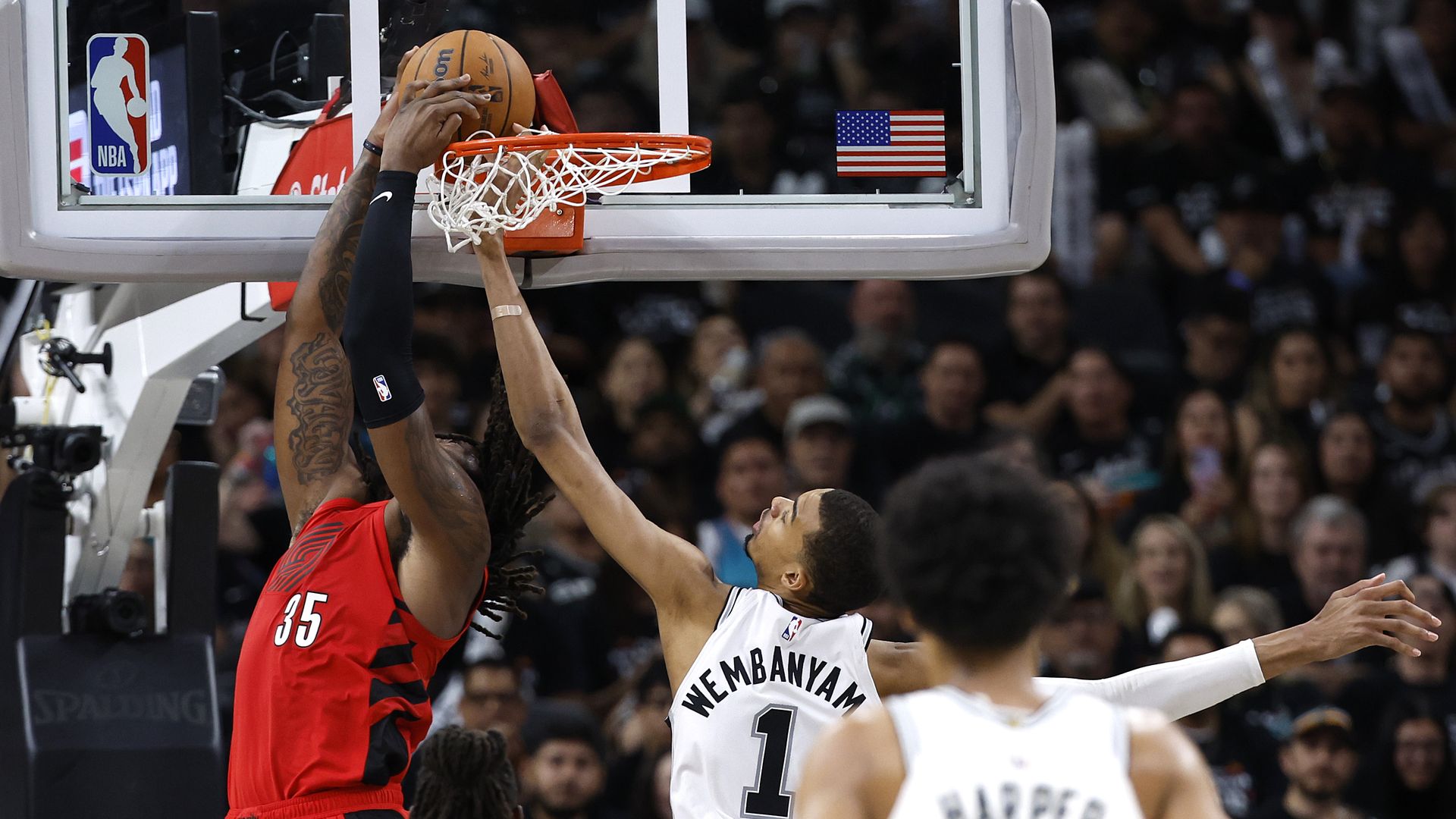 Two basketball players clash at the rim: a defender in red reaches up while an attacker in white jumps for the shot. An NBA logo and a flag sit on the backboard as a crowded arena watches.