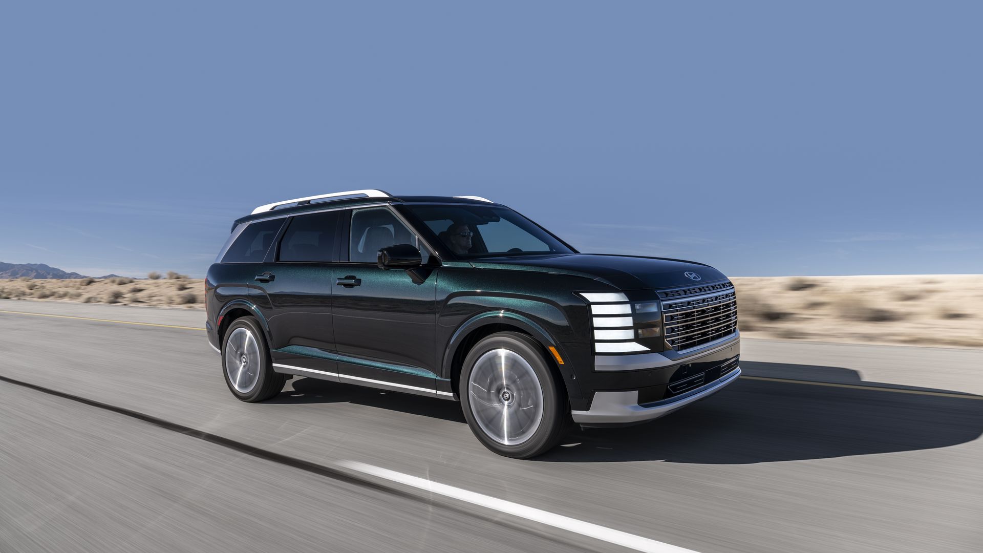 Dark green 2026 Hyundai Palisade SUV driving on highway with desert landscape and blue sky in the background.
