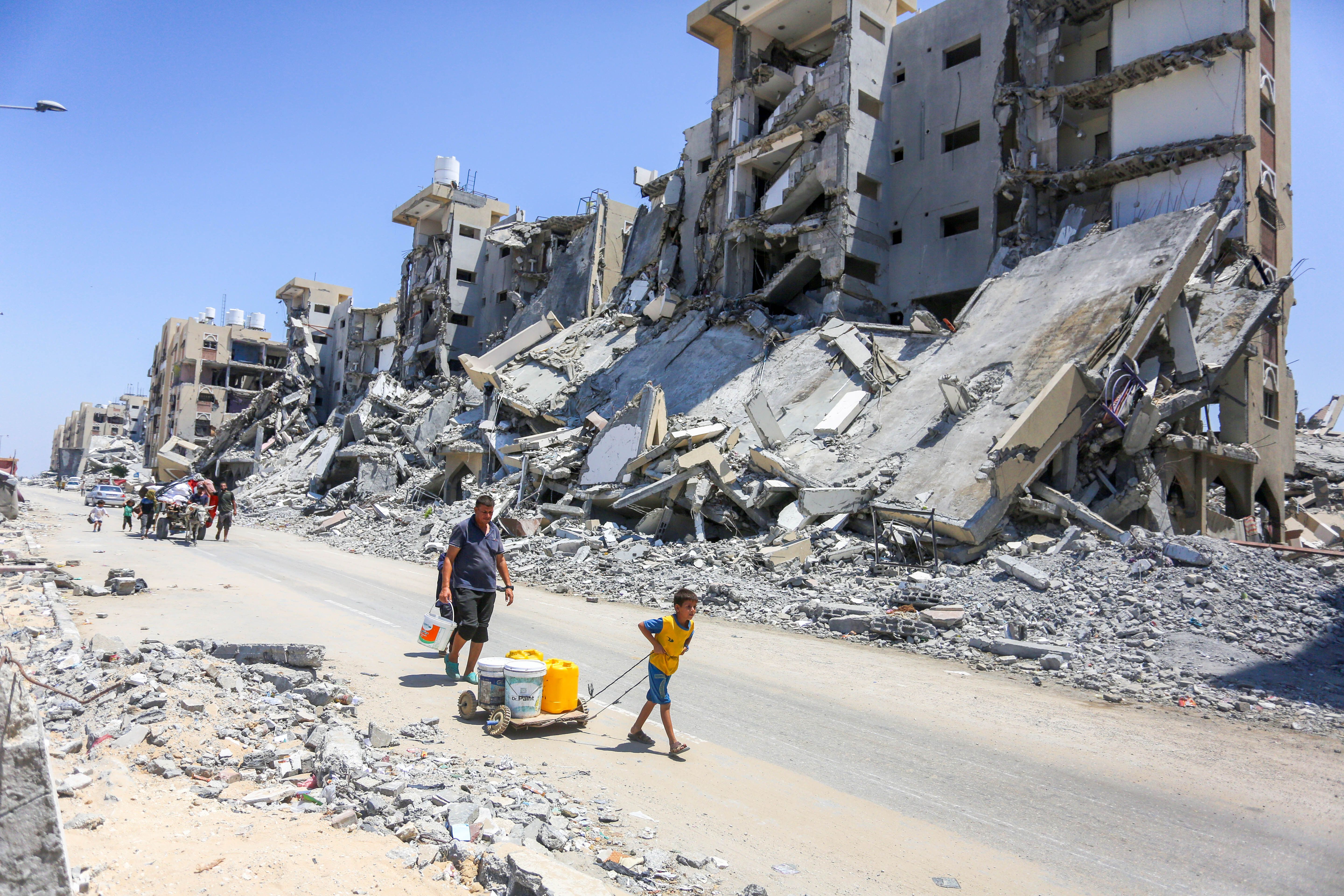A man and a child walk down a road flanked by destroyed buildings