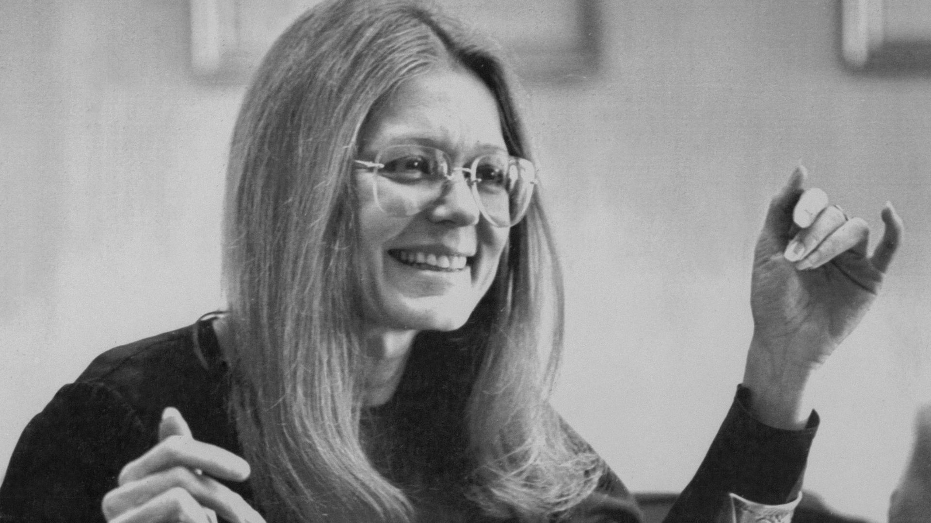 Black-and-white photo of Gloria Steinem smiling, with long hair and round glasses, seated at a desk and raising one hand in a gesture.