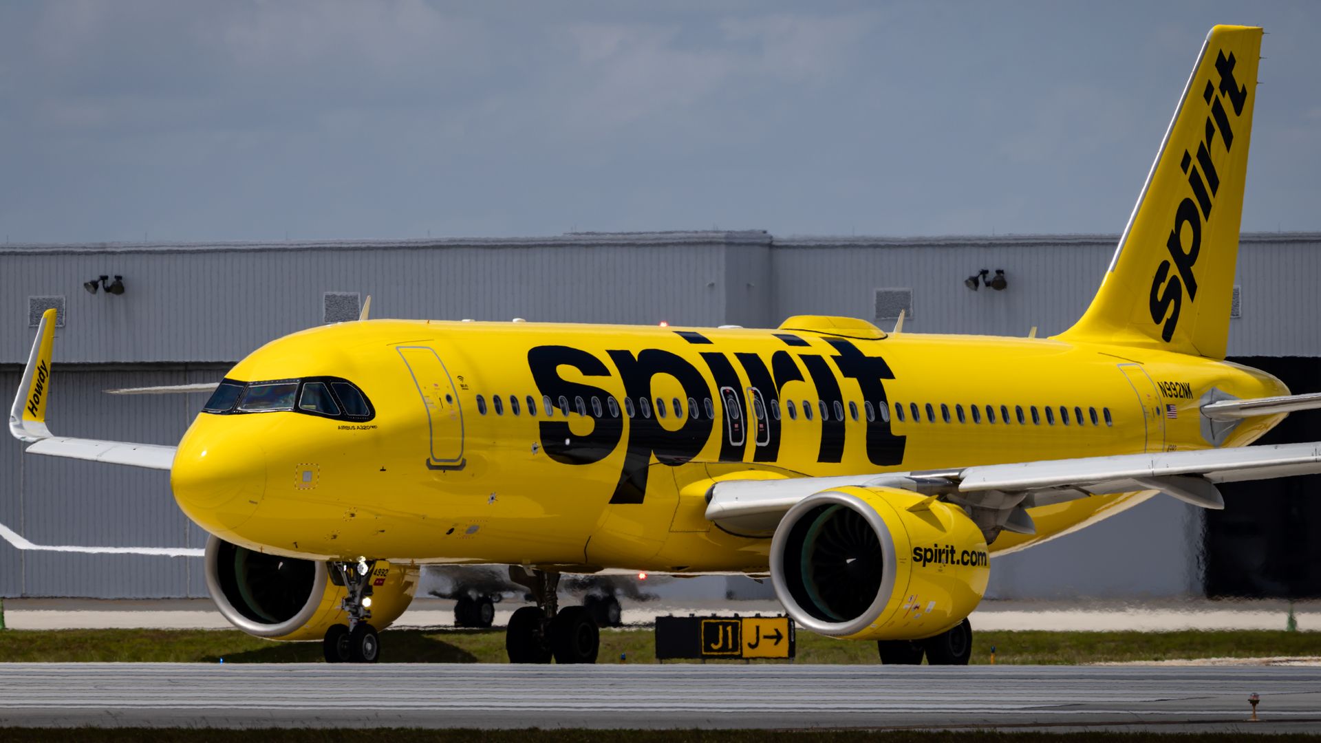 Bright yellow Spirit Airlines Airbus A320 parked on a tarmac beside a grey hangar; bold Spirit branding on the fuselage and vertical tail against a blue sky.