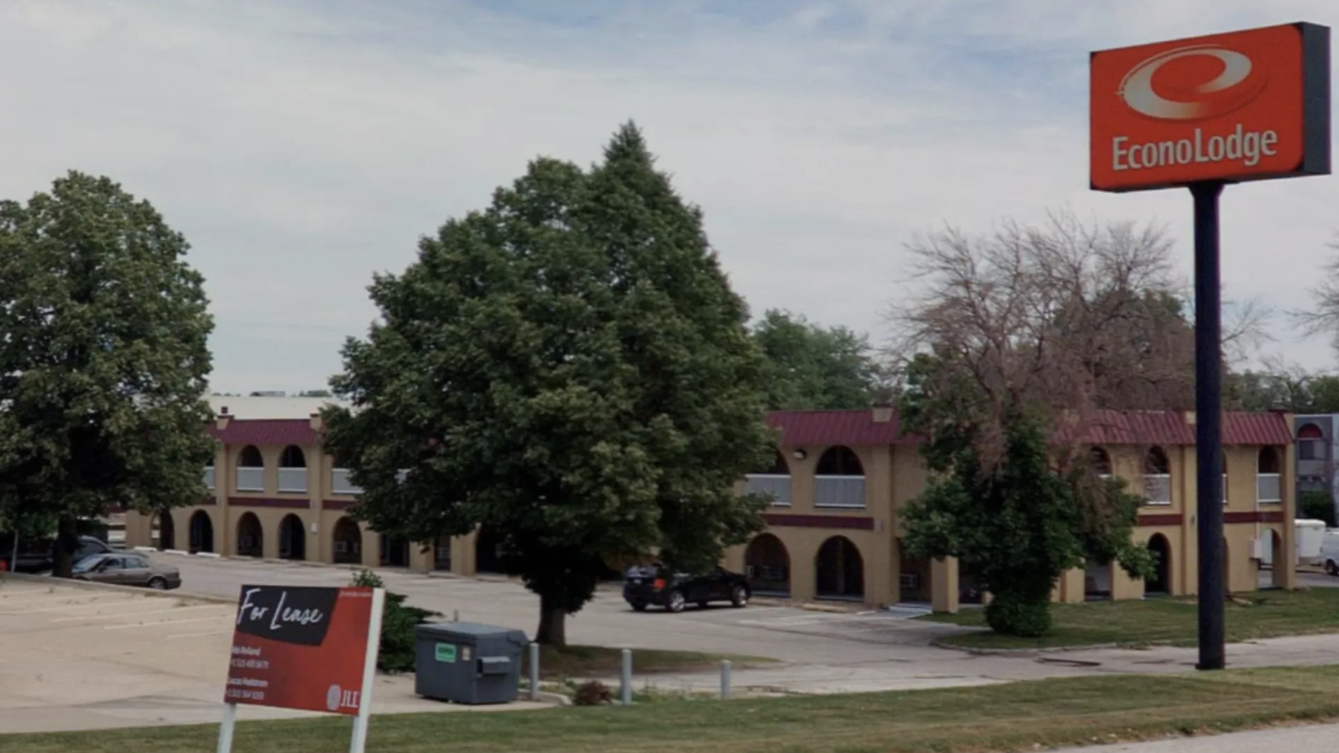 Exterior view of an Econo Lodge motel with beige walls and maroon trim, surrounded by green trees and a parking lot, under a cloudy sky.