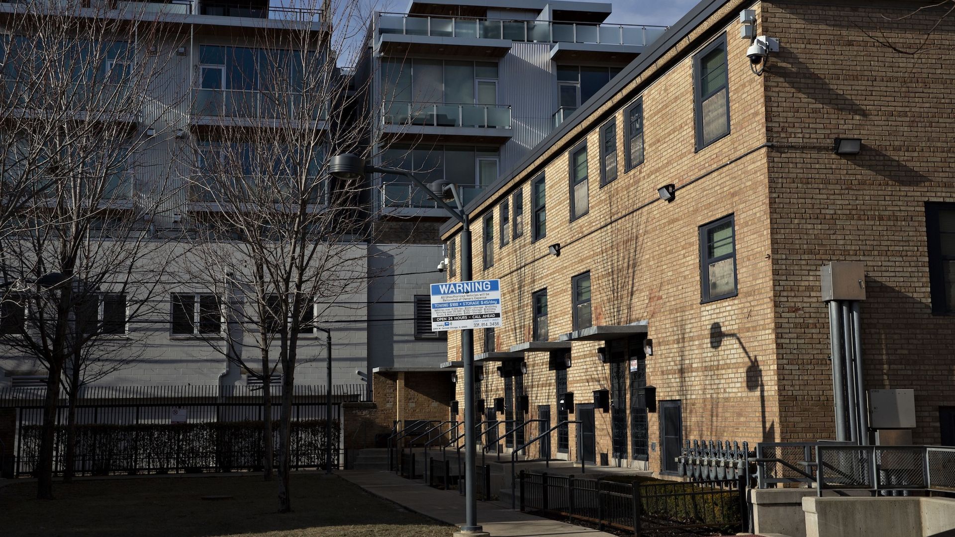 Red brick building with multiple doors and windows under a blue sky, leafless trees in front, and a warning sign on a lamppost in a fenced area with modern buildings behind in front of new modern condos.