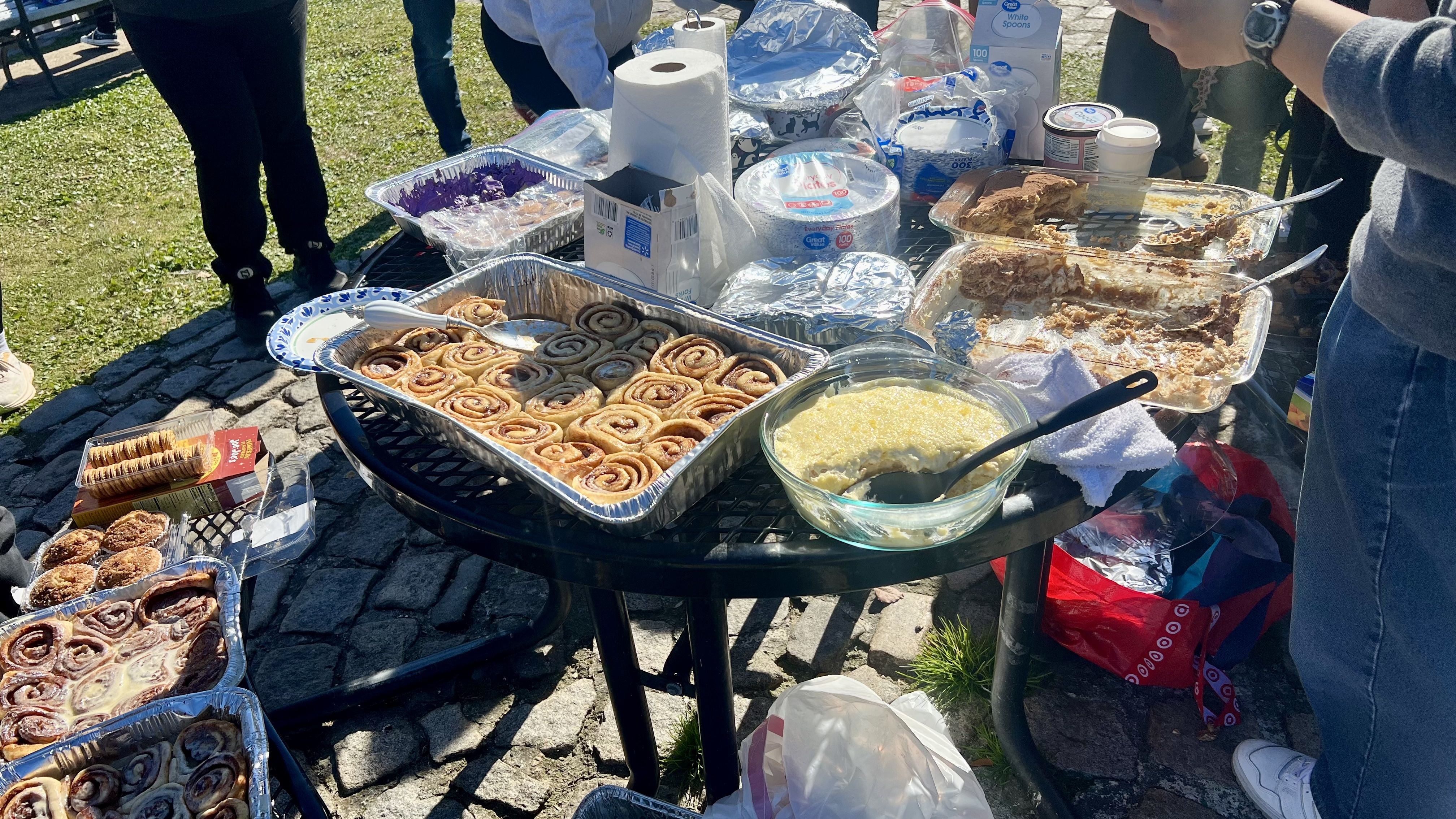 Outdoor table with trays of cinnamon rolls, bowls of creamy dessert, paper towels, disposable plates, and packaged food items, with people standing nearby on cobblestones and grass.