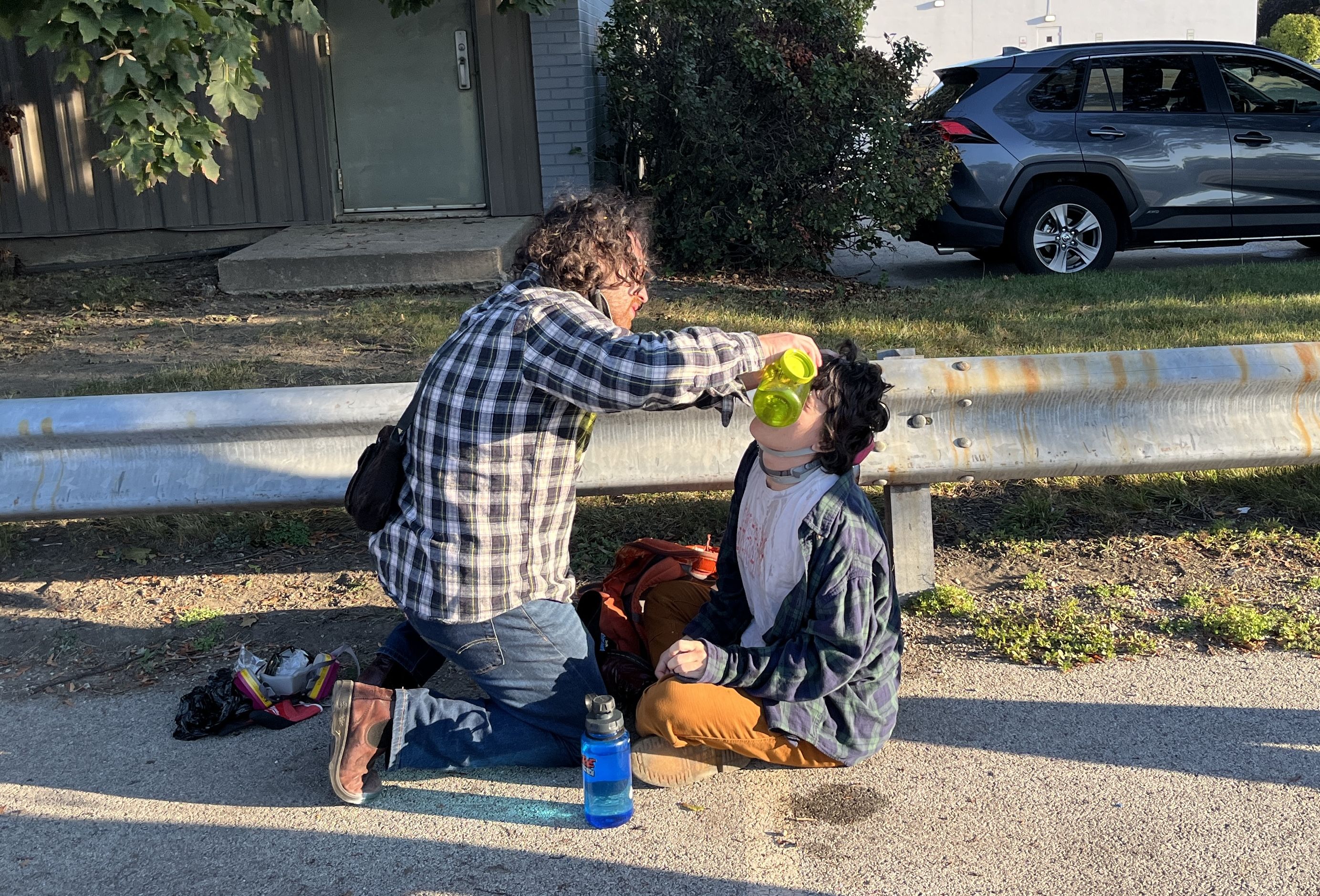 Two people outdoors next to a metal guardrail; one kneeling pours from a yellow bottle into the other's mouth, who is seated on pavement wearing a neck brace and flannel shirts.