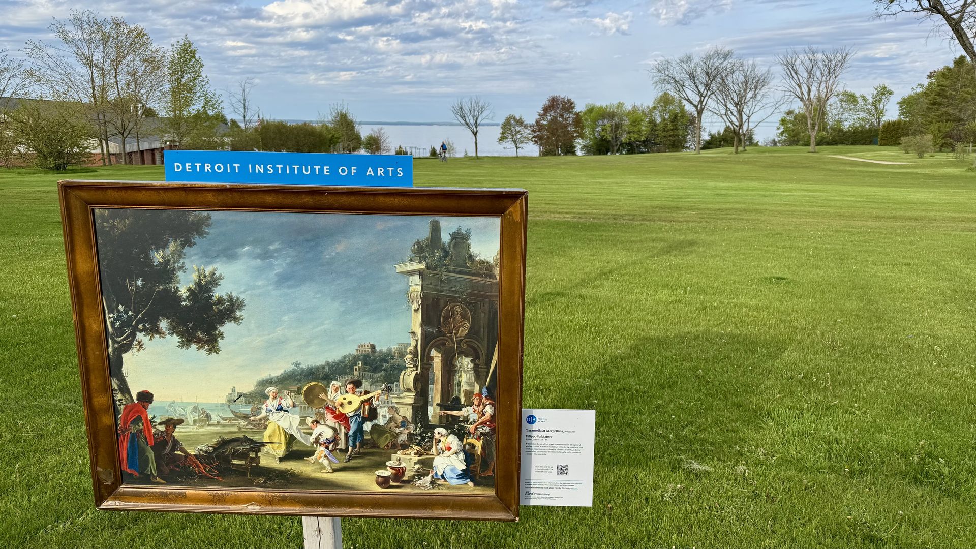 A painting framed and hammered into the ground on a stake in front of a giant lawn on Mackinac, with lakewater visible in the background