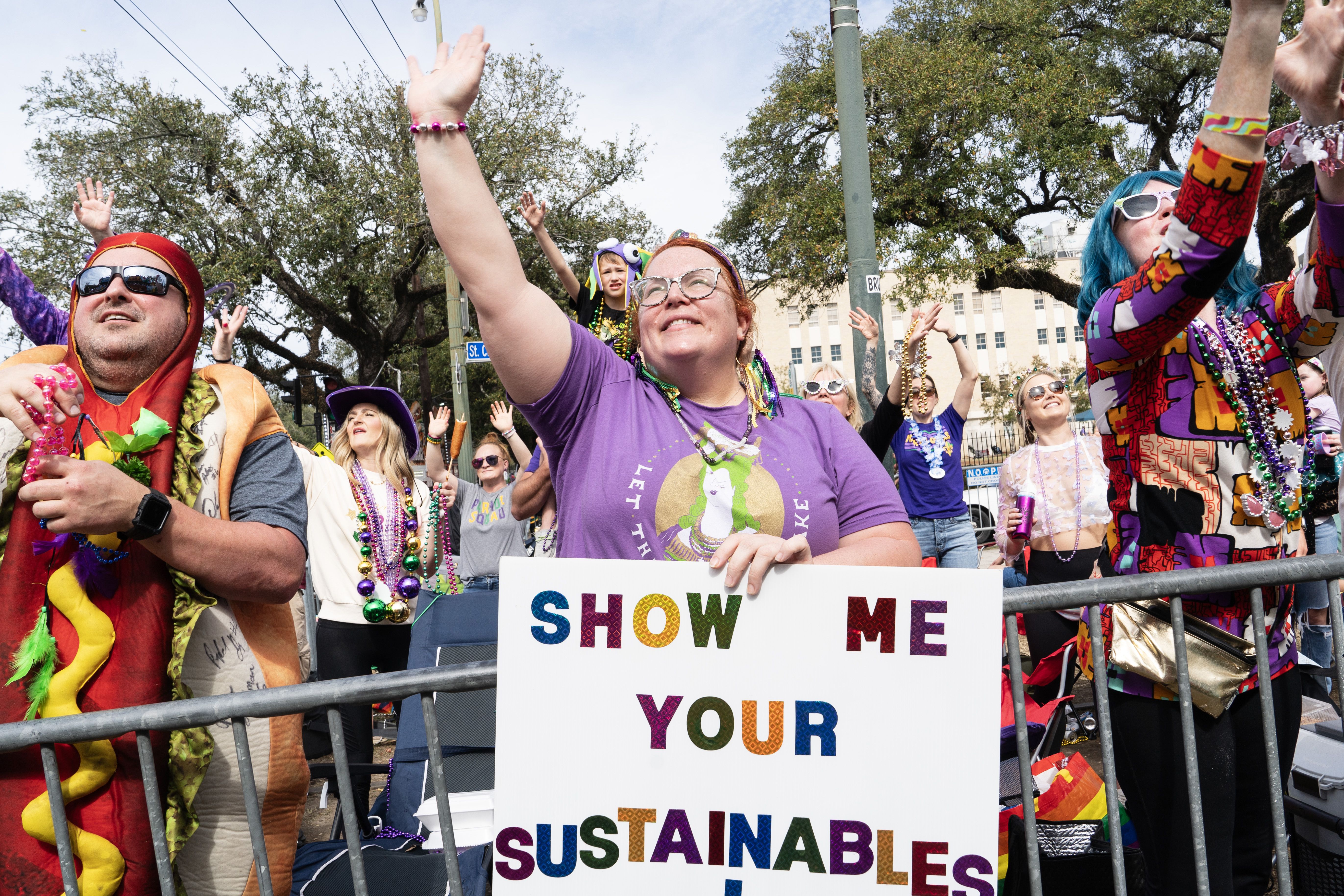 A woman tries to catch Mardi Gras throws as she stands at a barricade. She holds a poster that says "Show me your sustainables."