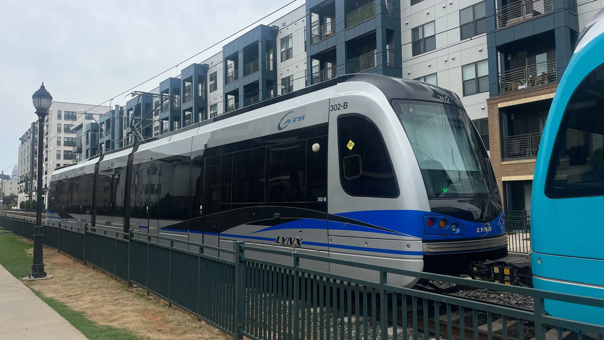 Modern LYNX light rail train in silver, black, and blue parked on tracks next to urban apartment buildings with blue and gray accents under a cloudy sky.
