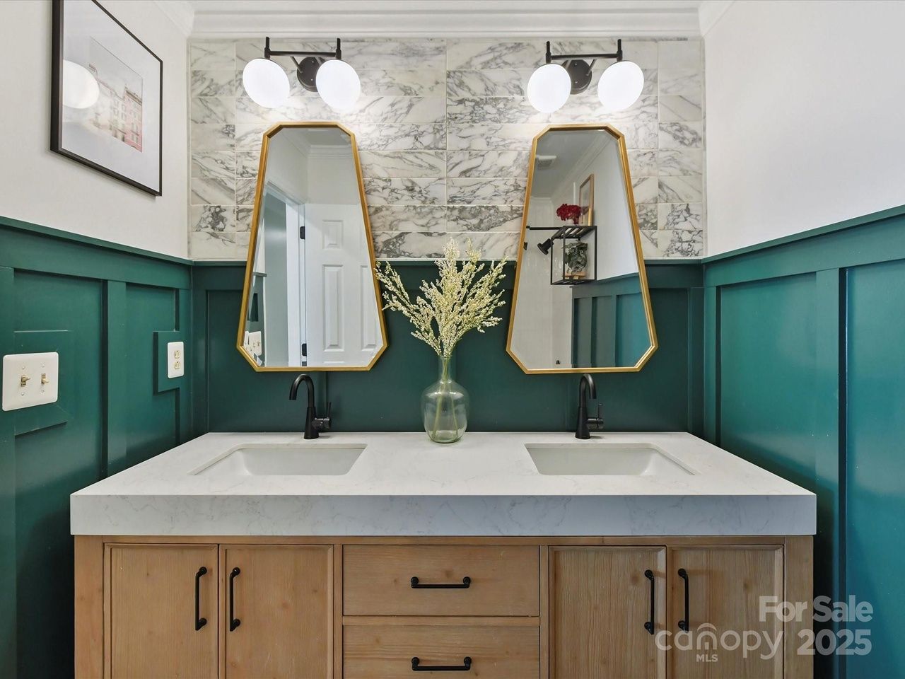Double sink bathroom vanity with wood cabinetry, teal panel walls, gold-framed mirrors, marble backsplash, black fixtures, and a glass vase with white flowers in the center under round white wall lights.