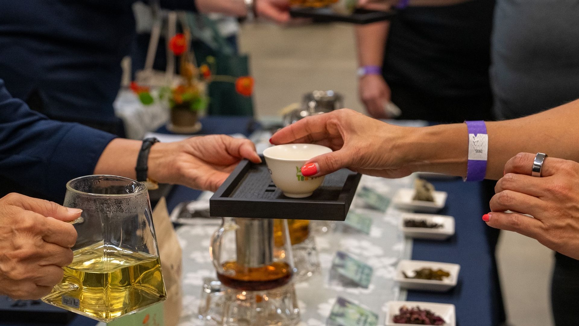 A person takes a cup of tea from a tray over a table laden with leaves and teapots. 