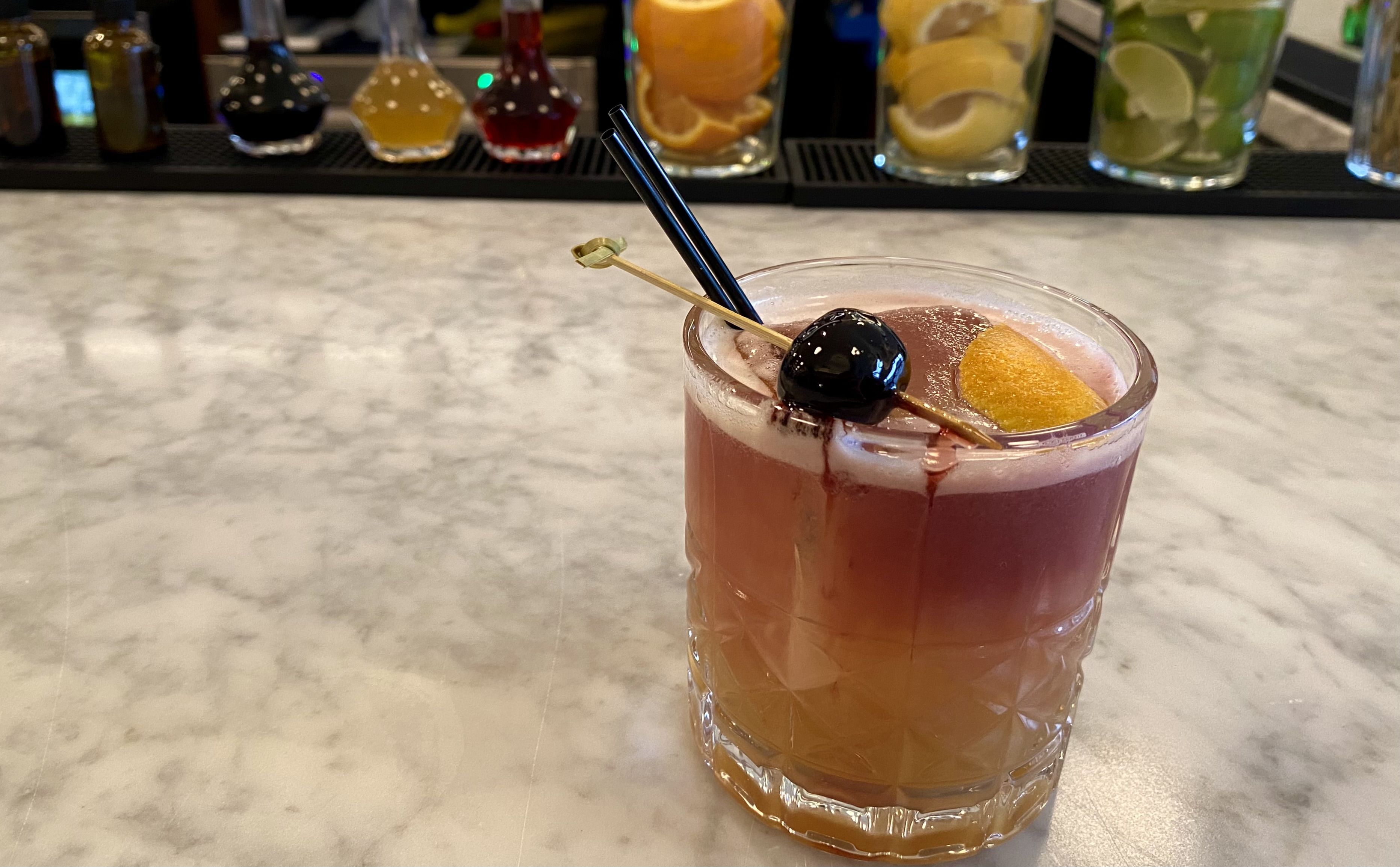 Layered cocktail in a short glass with amber and purple hues, garnished with a dark cherry and orange peel on a skewer, set on a marble bar counter with bottles and citrus slices in the background.