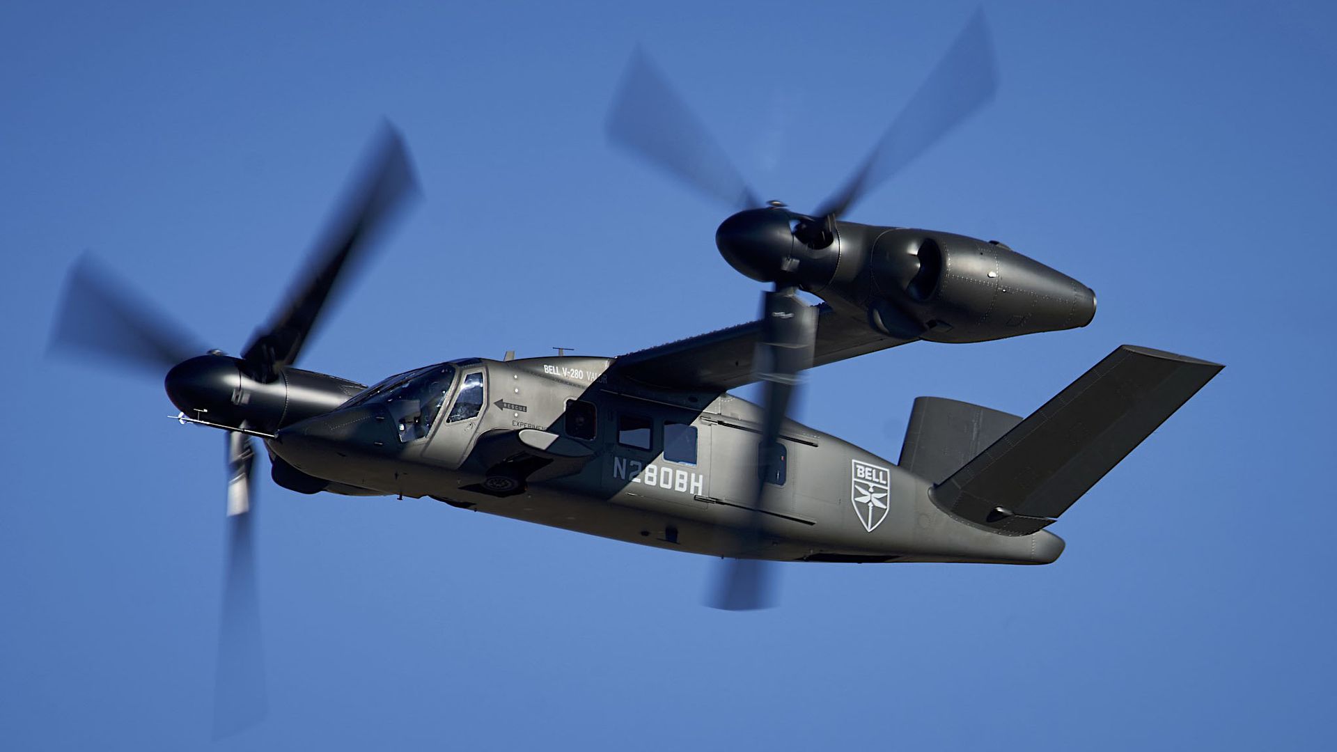 Gray Bell V-280 Valor tiltrotor aircraft in flight against a clear blue sky, showing spinning rotors and distinctive tail design.