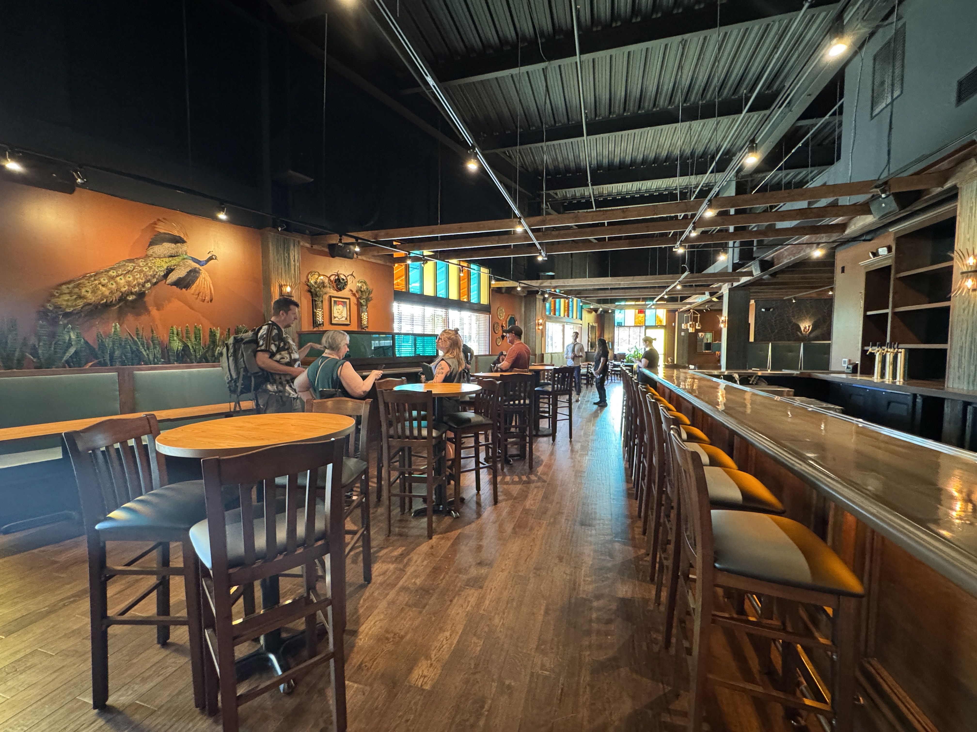 Interior of a cozy bar with wooden floors, high tables, and stools. Warm lighting, orange wall with a peacock mural, stained glass windows, and a few people standing and sitting near the tables.