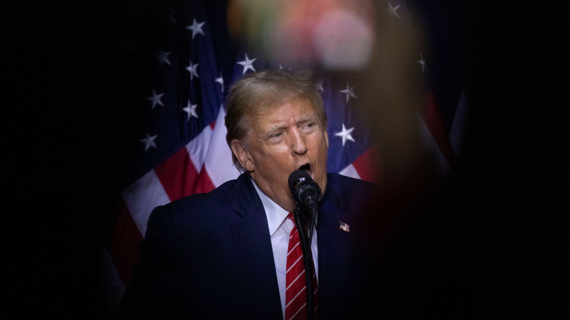 Former President Donald Trump, wearing a blue suit jacket, white shirt and red tie, standing in front of an American flag.
