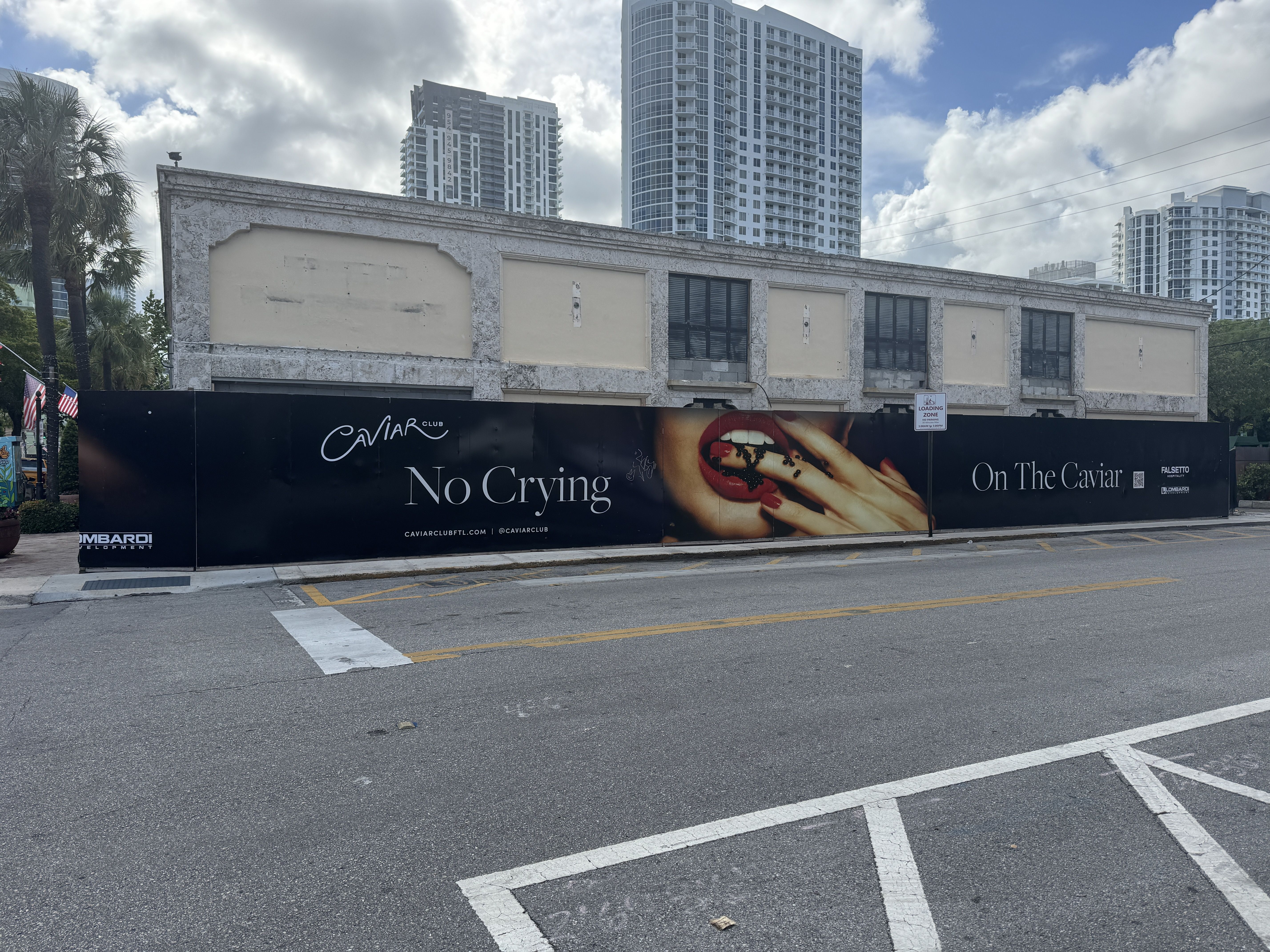 Street view of a storefront behind a long black fence advertising "No Crying" with red lips; white high-rises loom in the background under a blue, cloudy sky; palm trees on the left.