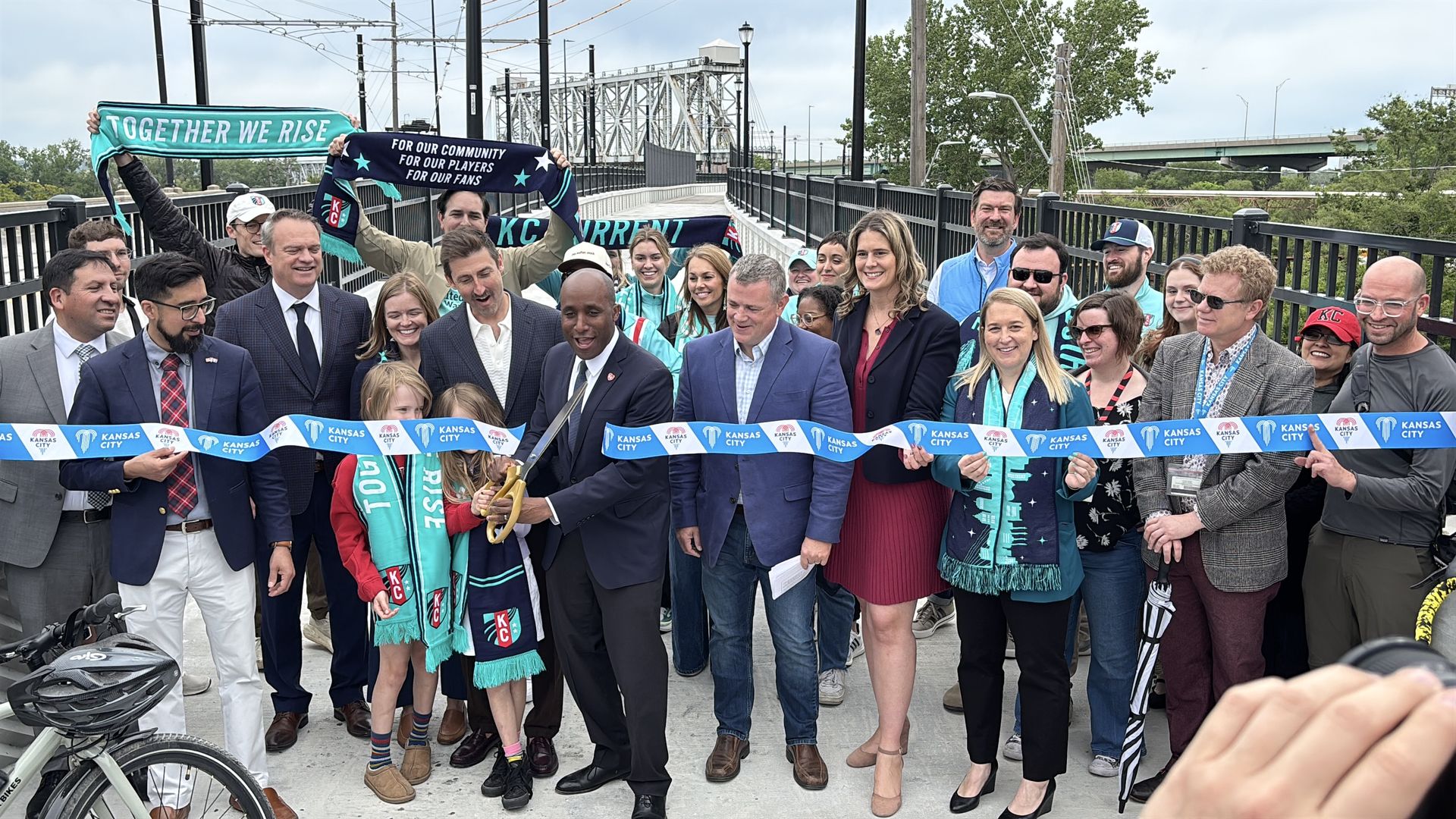 Diverse group on a bridge at a ribbon-cutting ceremony, holding a blue Kansas City ribbon and teal KC scarves. A man in a suit cuts the ribbon amid a crowd with a city bridge behind.