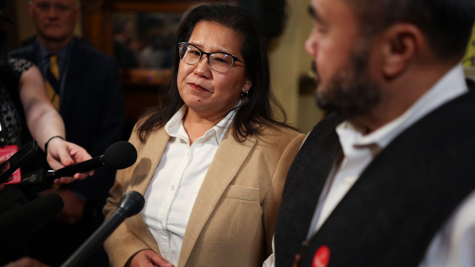 Woman in a tan blazer and white shirt wearing glasses speaks to reporters at a press event. She looks thoughtful as microphones are held up, with a suited man blurred in the background.