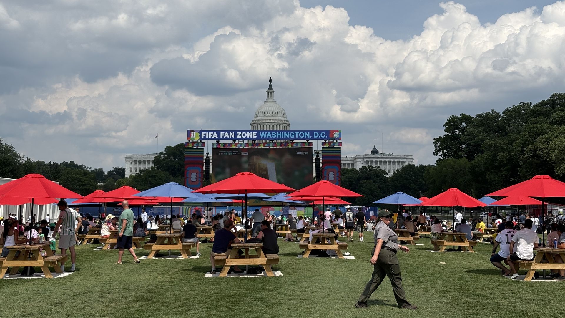 Busy outdoor FIFA Fan Zone in Washington, D.C. with red and blue umbrellas over picnic benches, a large stage labeled "FIFA FAN ZONE WASHINGTON, D.C.", and the Capitol dome against a cloudy sky.