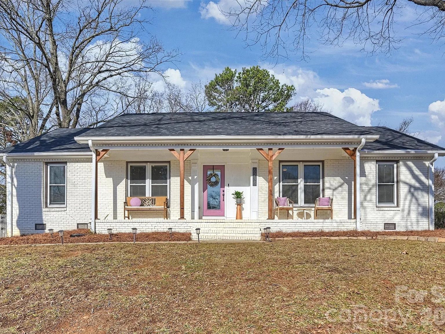 Single-story white brick house with black roof, pink front door, wooden porch posts, two chairs with pink cushions, and a bench with a pink pillow under a clear blue sky.