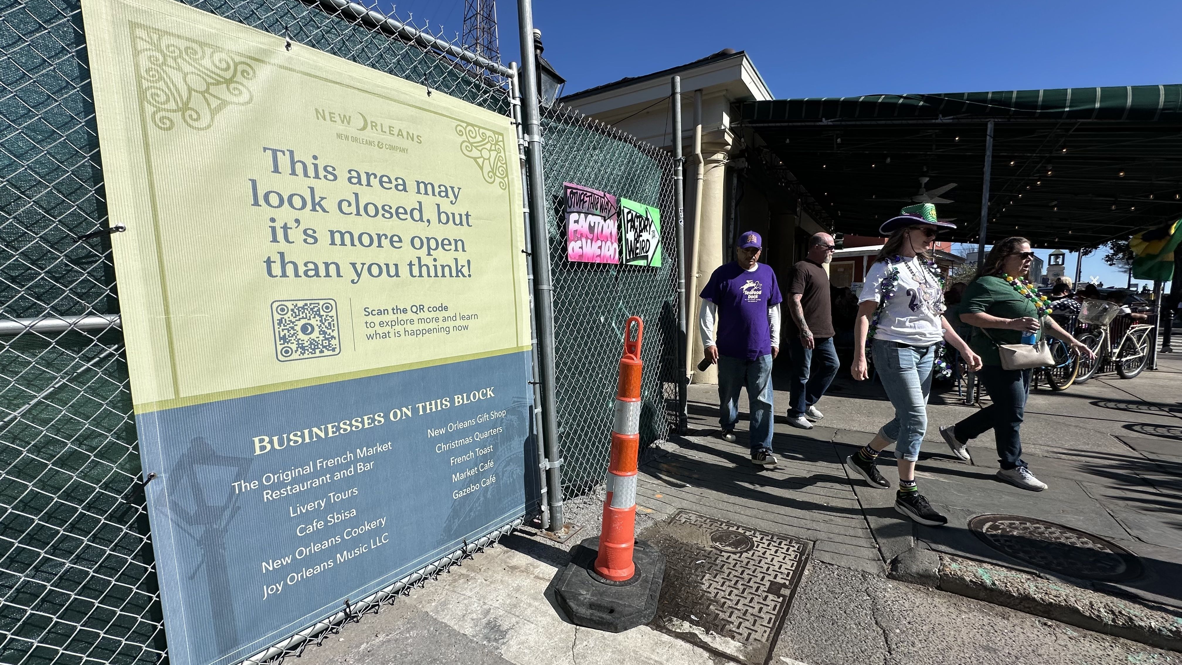 Yellow and blue sign on chain-link fence in New Orleans says the area looks closed but is open, with a QR code and list of businesses. People walk nearby under blue sky.