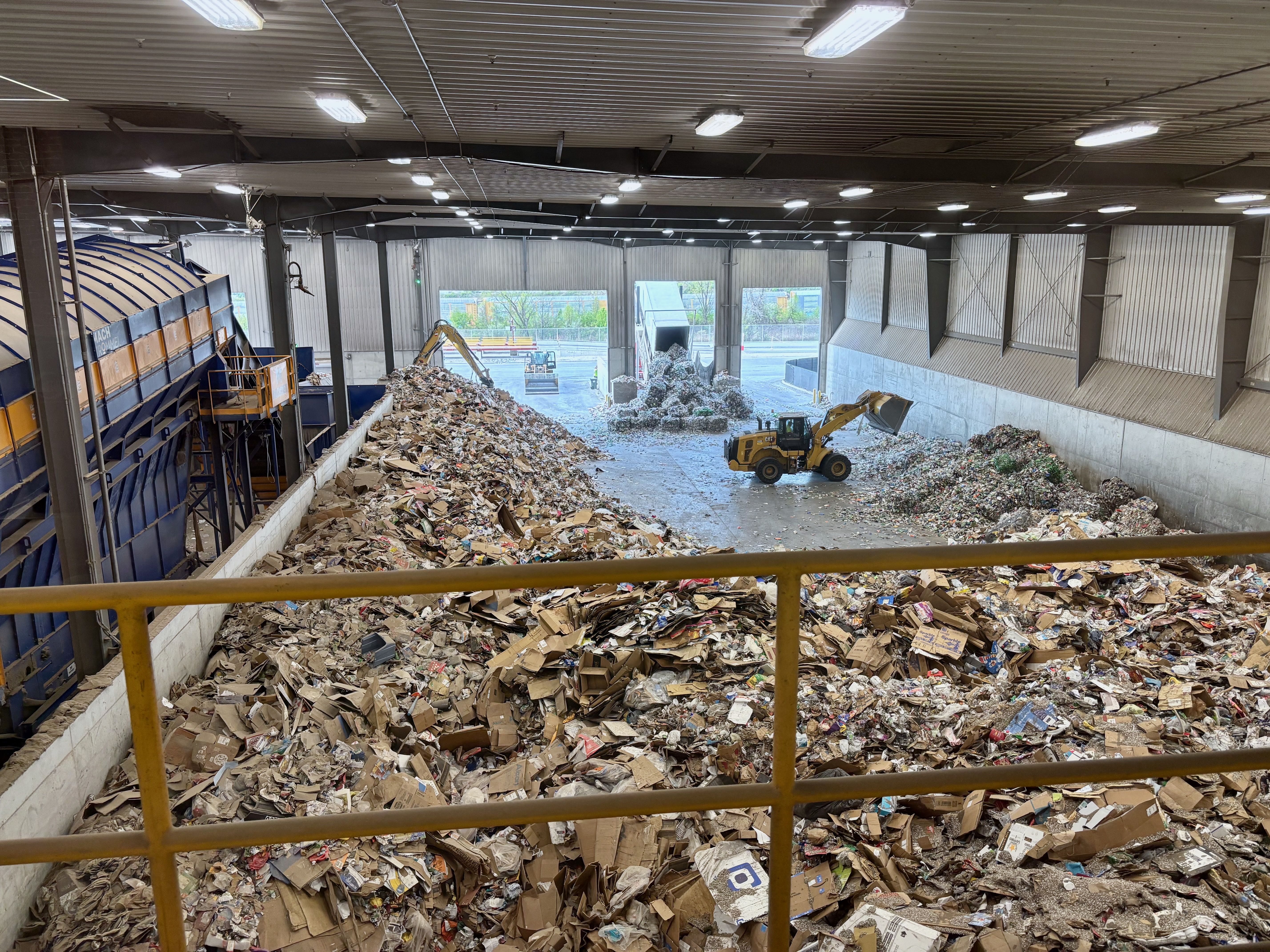 Inside a large industrial warehouse, massive piles of cardboard and debris cover the floor. A yellow front-end loader sits amid rubble near gray metal walls under bright overhead lights.