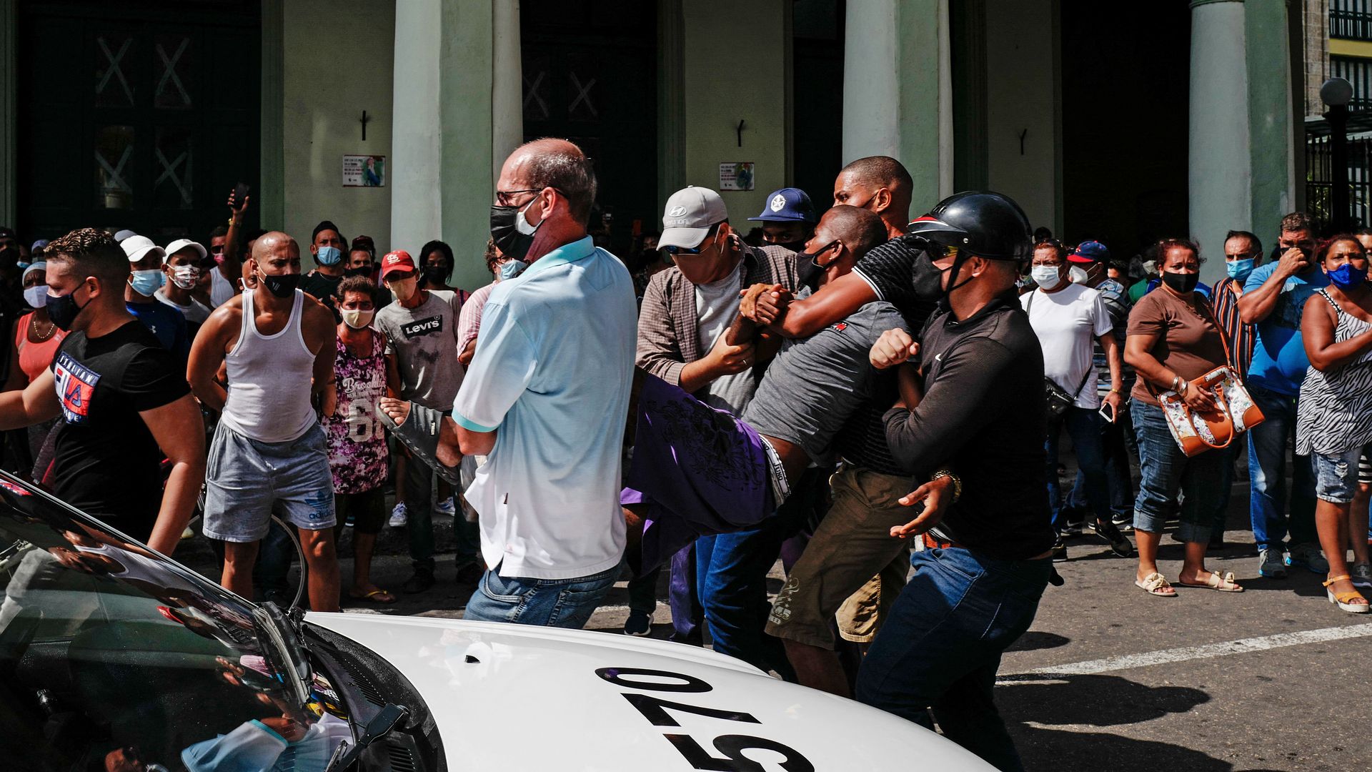 Photo of riot police carrying a detained protester to a police vehicle while other protesters watch from behind