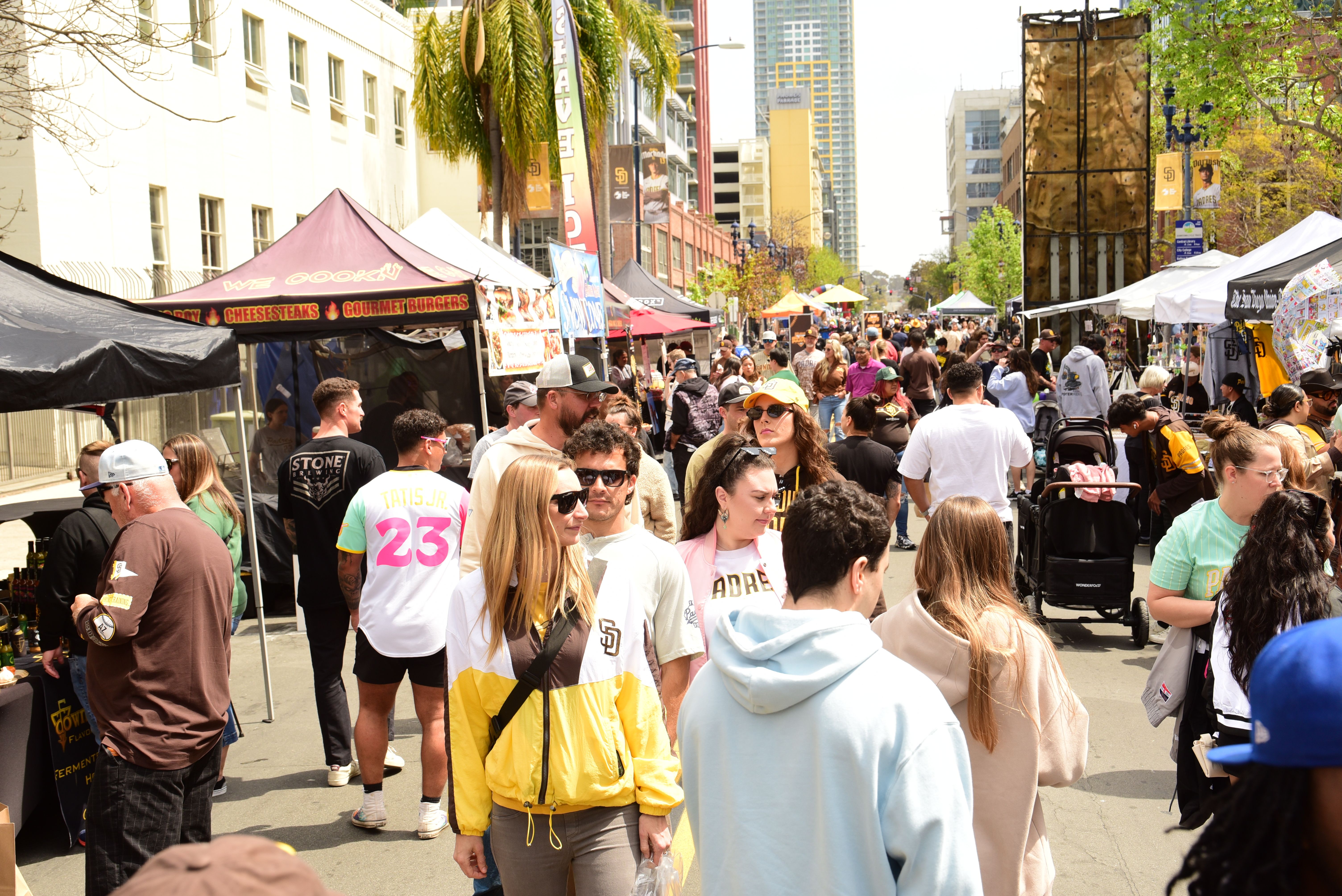 Crowded city block party with colorful vendor tents, Padres fans, shoppers, banners, and stalls; palm trees and tall buildings line the road in the background.
