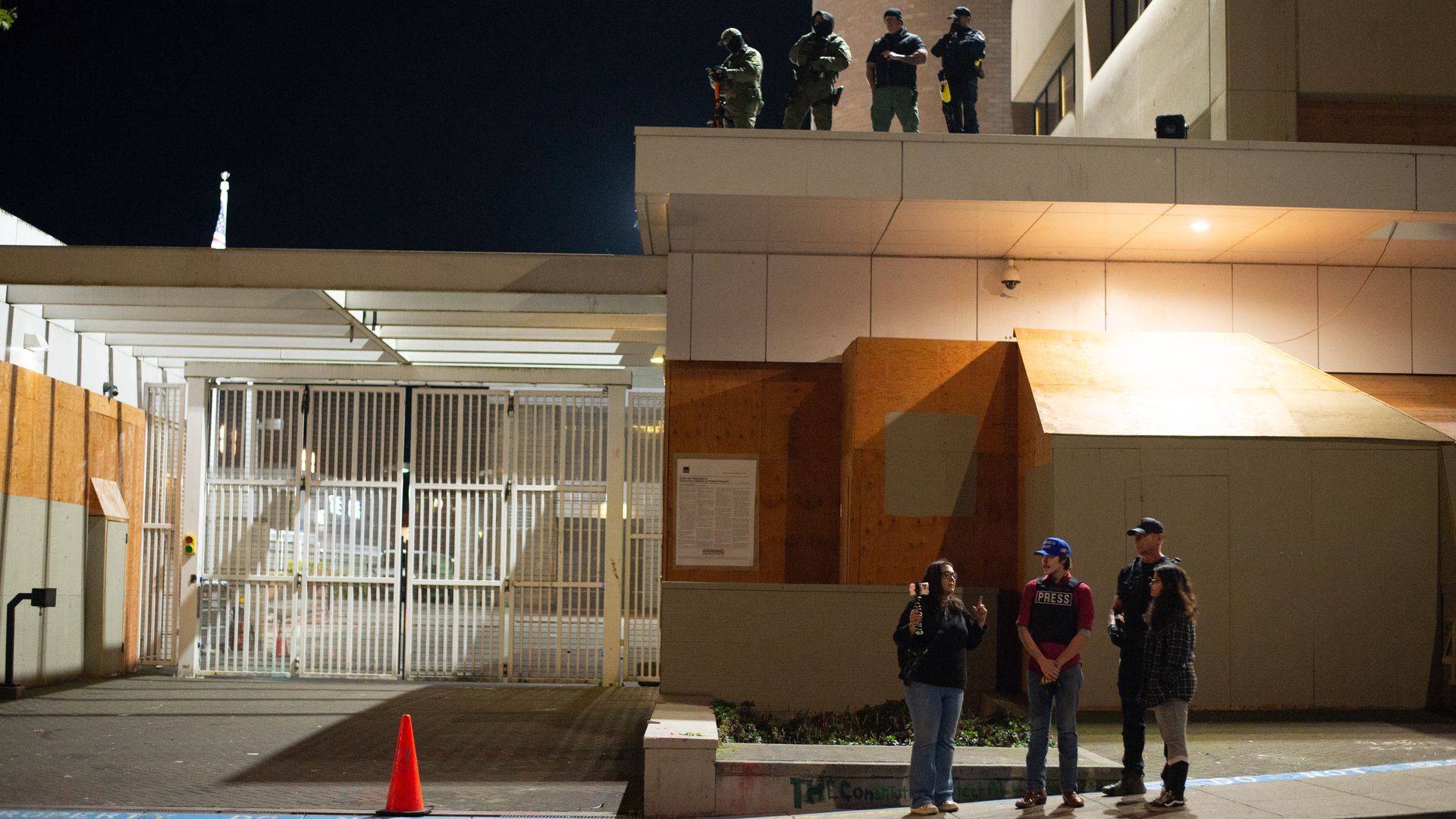 Night scene outside a building with boarded windows and a metal gate, four armed security personnel stand on the rooftop, while four civilians, including one with a press vest, stand on the sidewalk.