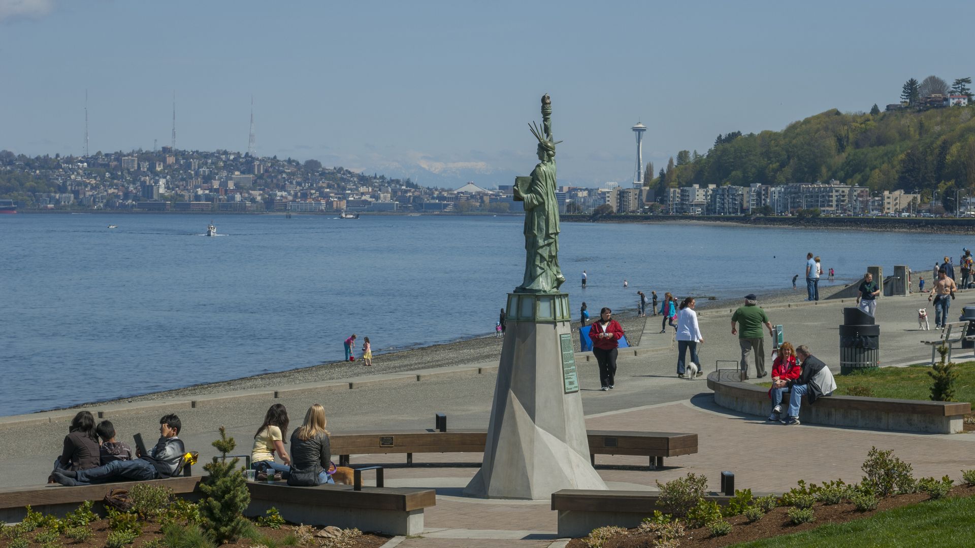 Outdoor scene by waterfront with a small Statue of Liberty replica on a pedestal, people walking and sitting on benches, a blue sky and cityscape including the Space Needle in the background.