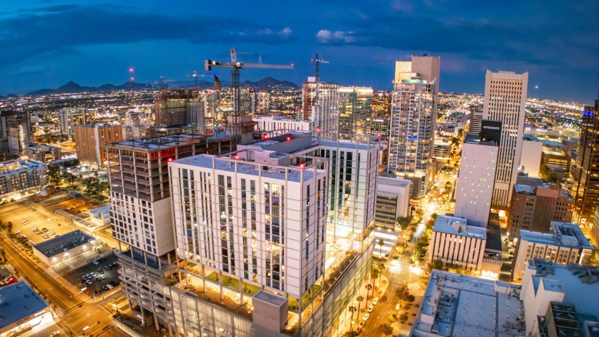Aerial view of downtown Phoenix at dusk, featuring illuminated high-rise buildings and active construction cranes.