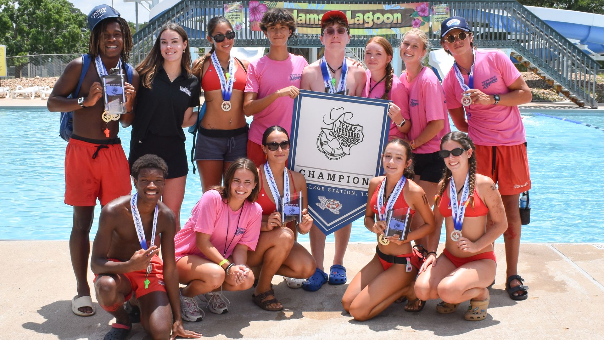 Group of smiling lifeguards posing with medals and a championship plaque by a pool at a water park; large blue water slide and an American flag in the background.